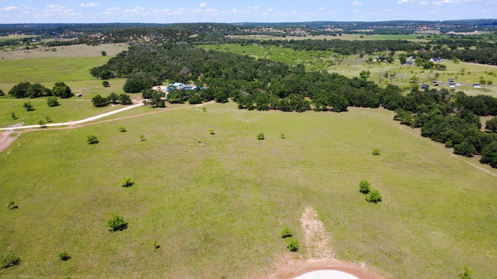 A vast open field with scattered trees and a dirt road, leading to a cluster of buildings with blue roofs surrounded by greenery, under a partly cloudy sky.