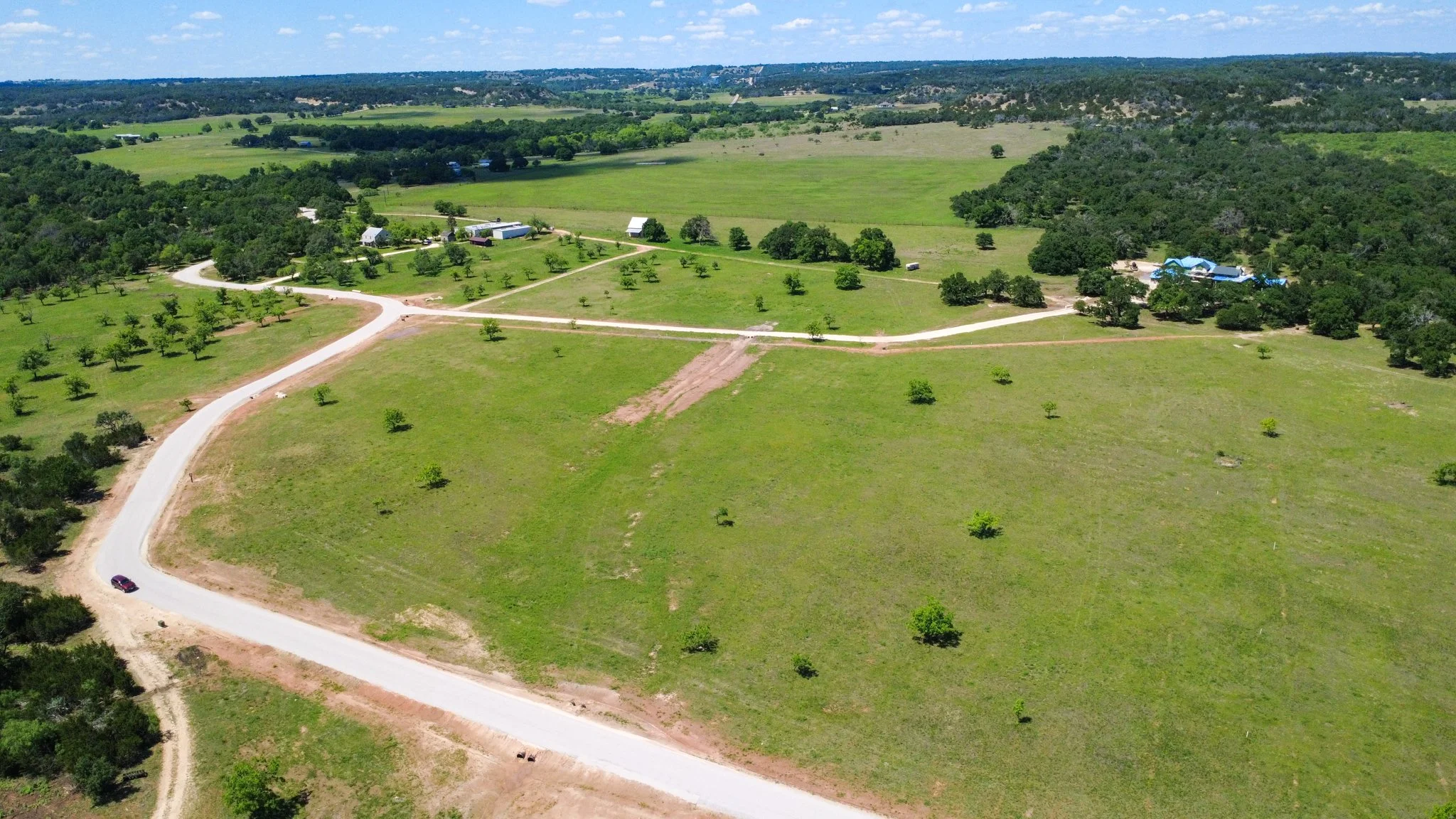 Aerial view of a rural area with green fields, trees, dirt roads, and a few buildings with blue roofs in the distance.