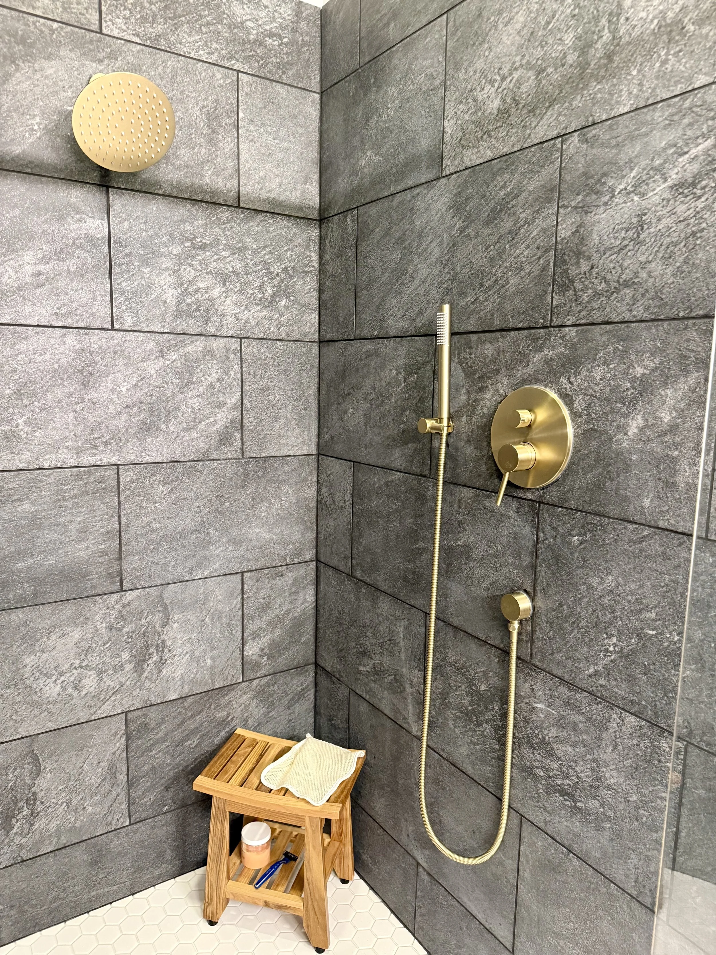 A shower area with dark gray tiled walls and white hexagonal tiled floor. There is a yellow showerhead, a gold shower control, and a gold handheld shower. A small wooden stool with a towel, a cup, and toiletries is in the corner.