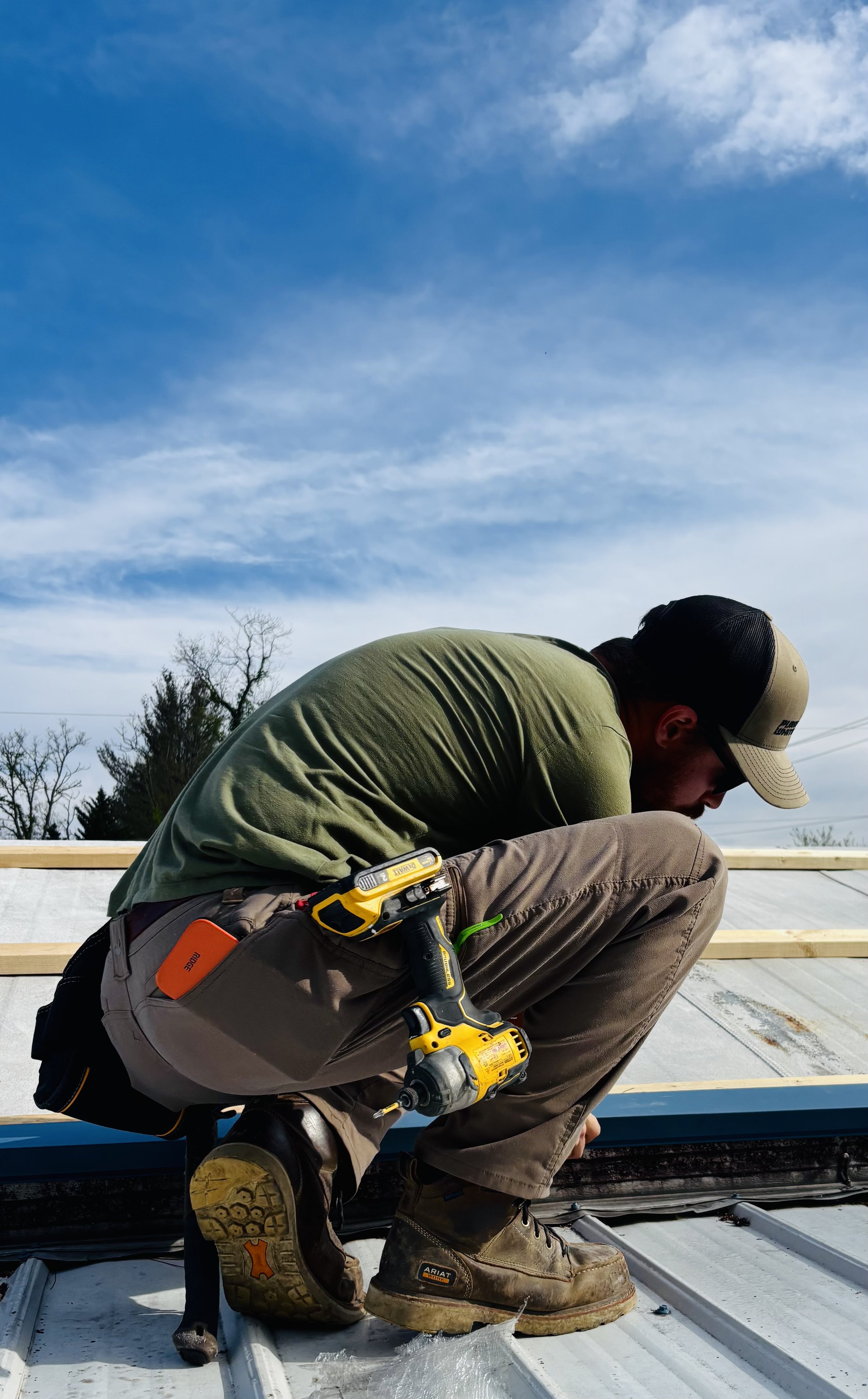 A man wearing a cap, green shirt, and brown work pants kneels on a roof during daytime, using a yellow cordless drill with a black and yellow tool belt. He is working with wooden framing on a flat, partially covered surface, with leafless trees and a cloudy sky in the background.