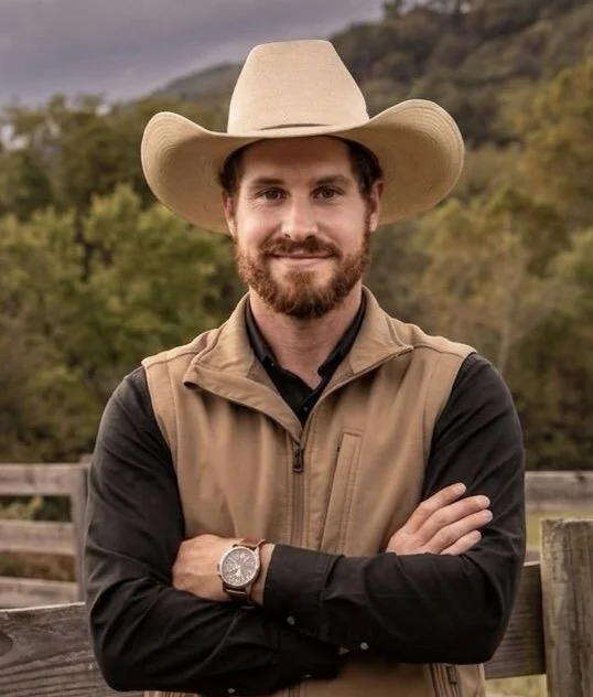 Man wearing a cowboy hat and vest, smiling with arms crossed outdoors.