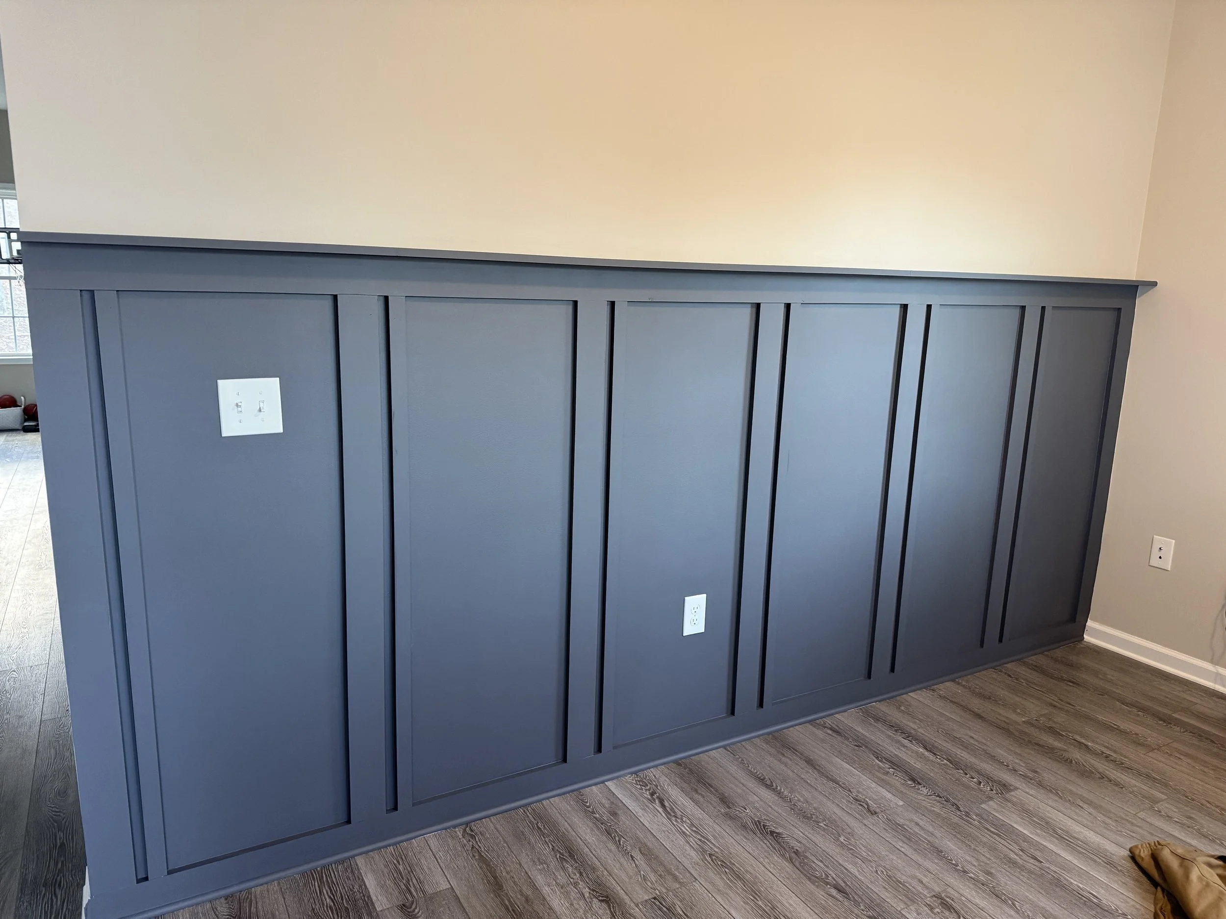 A dark blue wooden headboard with vertical panel details against a beige wall. There are electrical outlets on the headboard and a hardwood floor underneath.