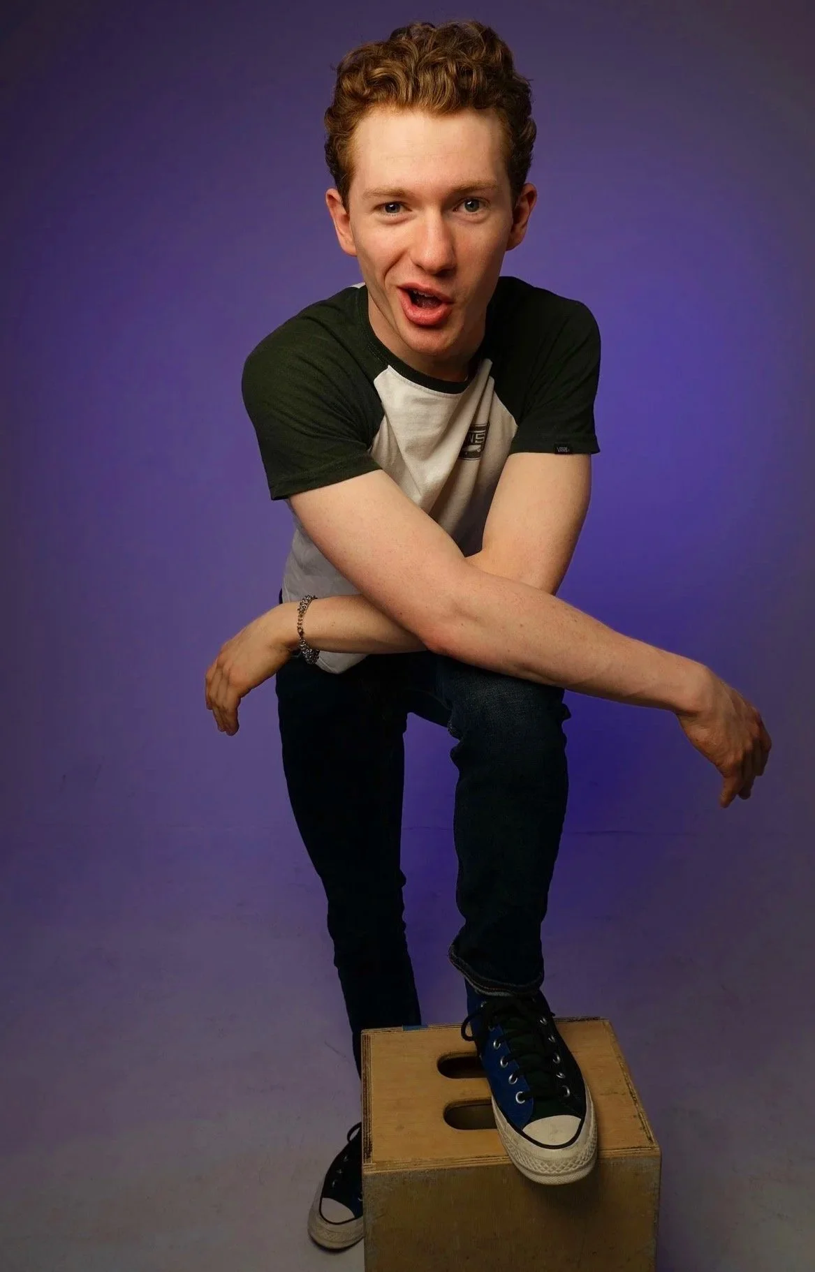 A young man with curly red hair wearing a black and white T-shirt, standing on a wooden box and balancing with one foot on the box, against a purple background.