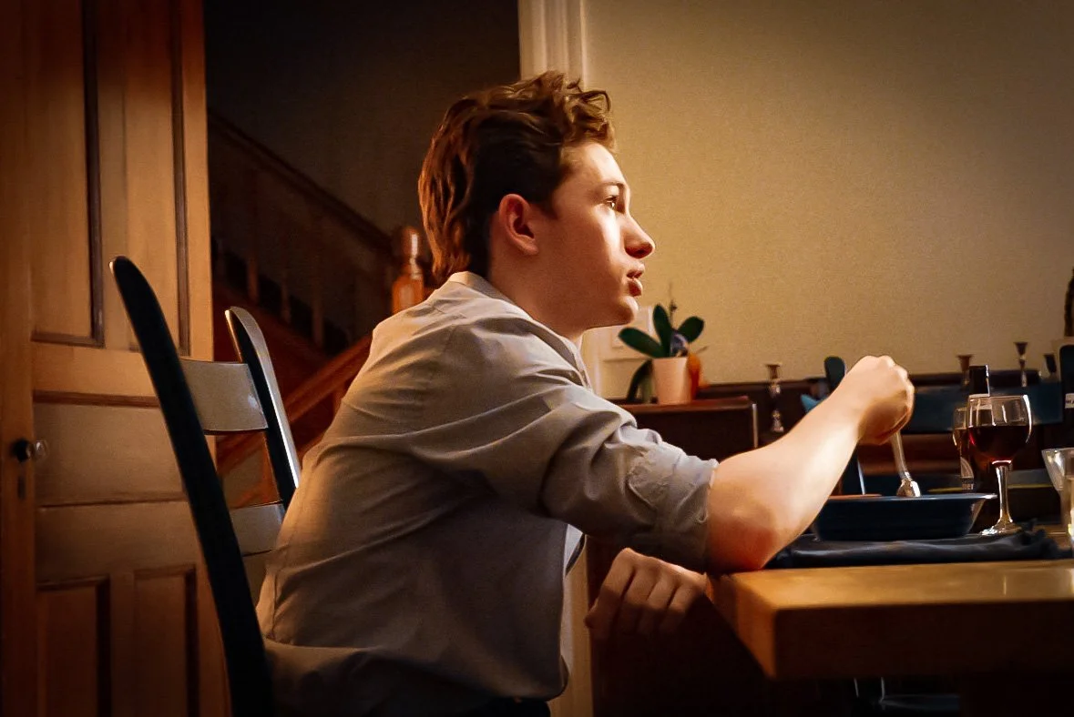 Young man sitting at a dining table with a glass of red wine, eating with a fork, in a warmly lit room.