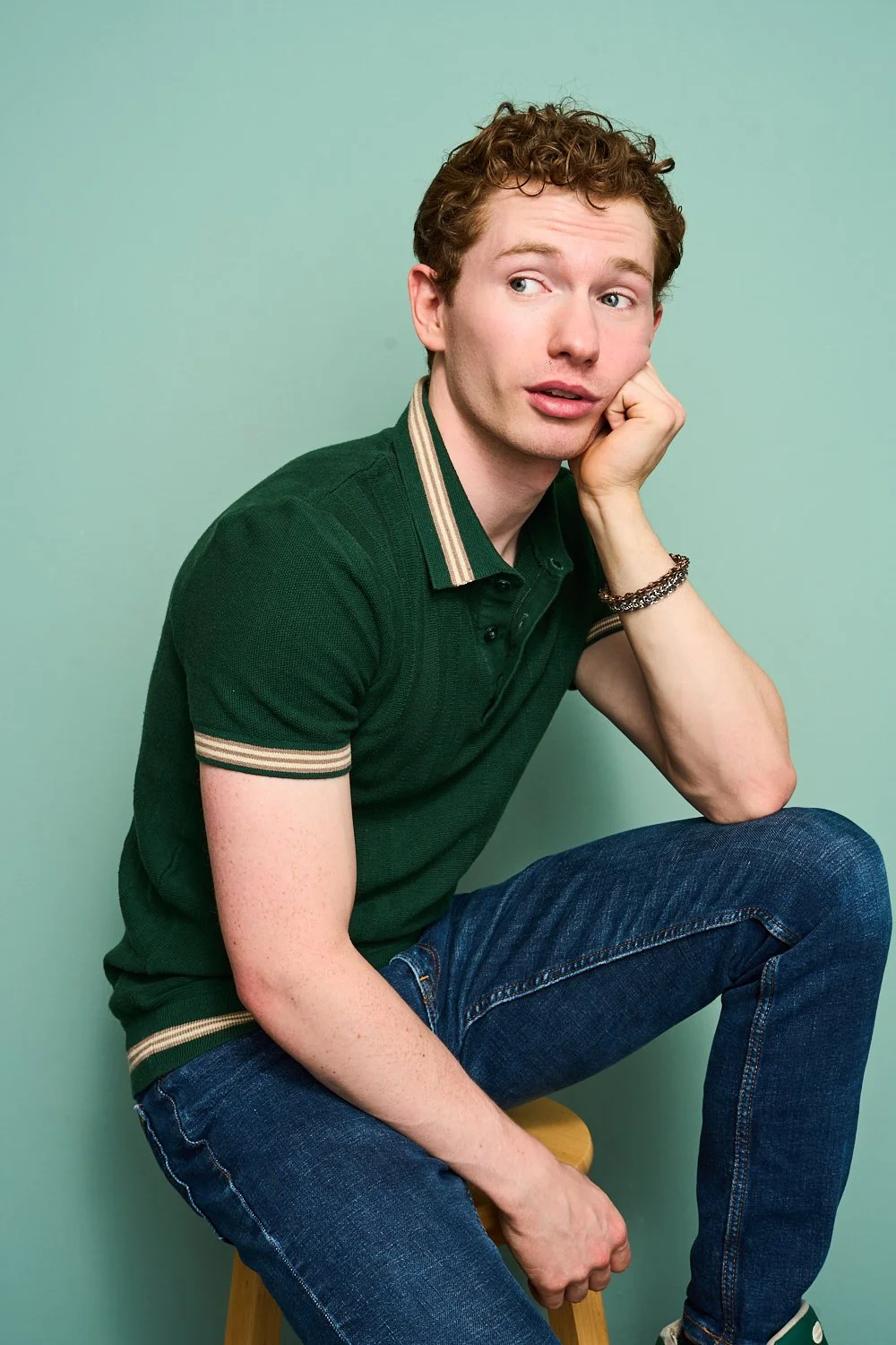 A young man with curly hair wearing a dark green polo shirt and blue jeans, sitting on a wooden stool against a light green background, resting his face on his hand and looking to the side.