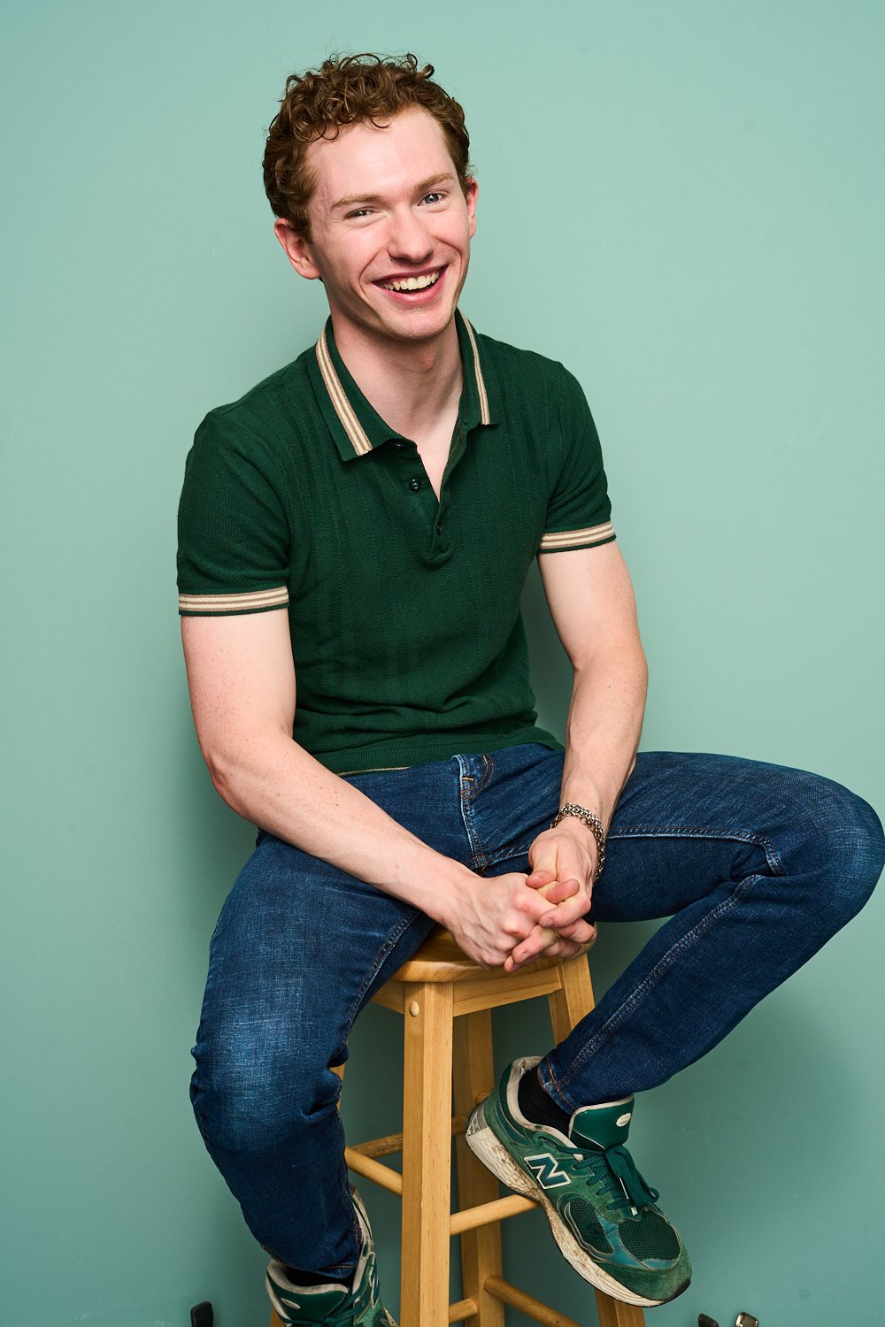A young man with curly hair, wearing a green polo shirt and jeans, sitting on a wooden stool against a plain light green background, smiling and looking at the camera.