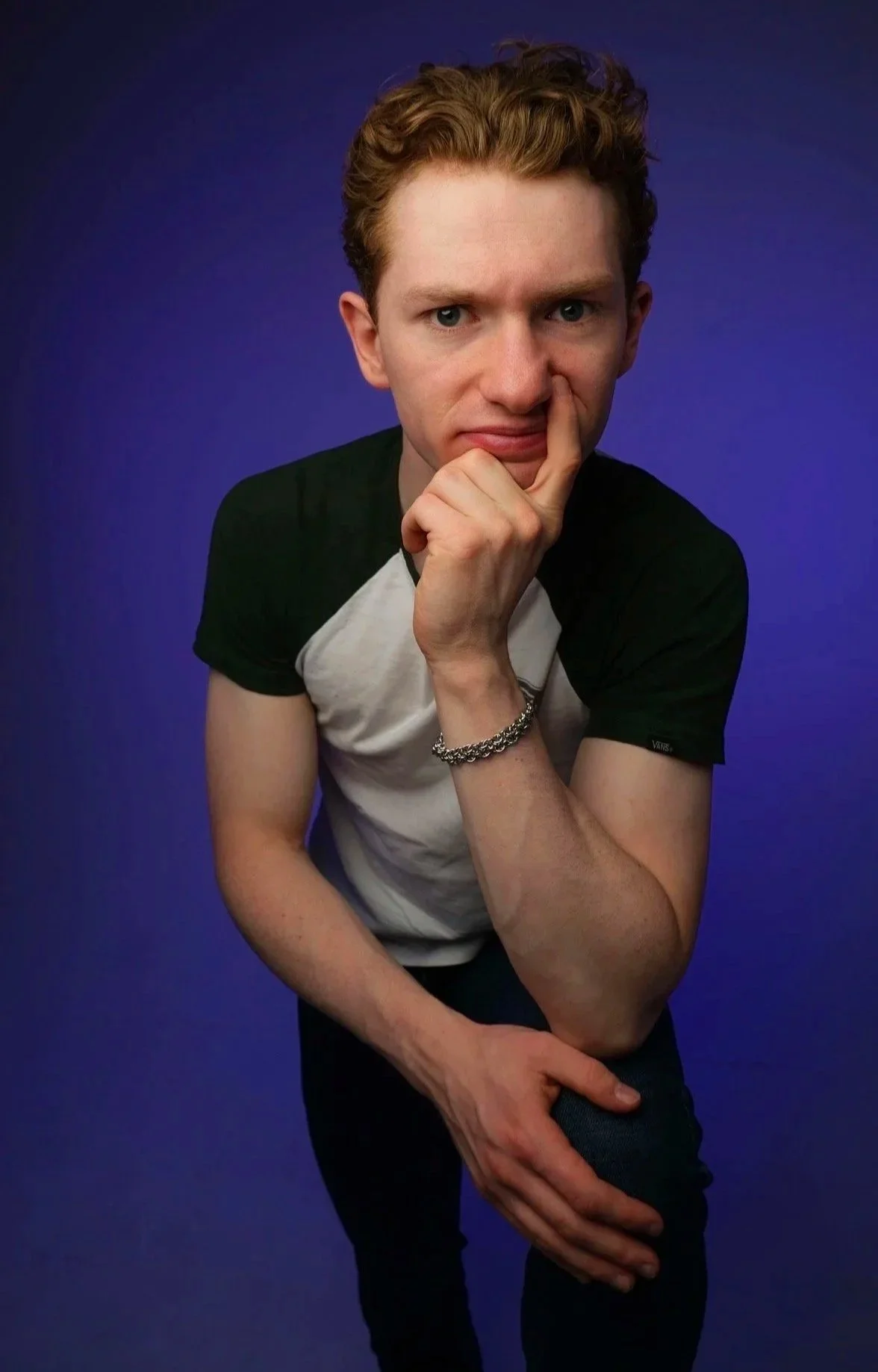 A young man with curly red hair and blue eyes posing against a dark blue background. He is wearing a black and white t-shirt, a silver bracelet, and is touching his face with his right hand while slightly bending his left arm.