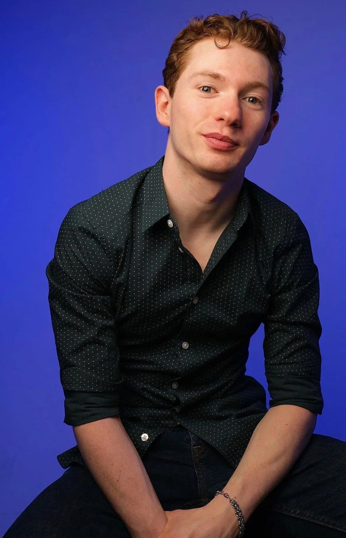 A young man with red hair and fair skin, wearing a black shirt with white polka dots, seated against a blue background, looking at the camera with a slight smile.