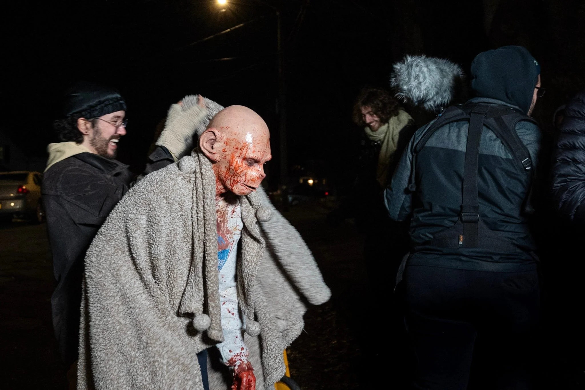 A person wearing a mask and gloves adjusts a life-sized Halloween costume of a bloody, wounded character with a bald head and tattered clothing. Two other people in winter clothing and hats observe the scene in a dark outdoor setting.