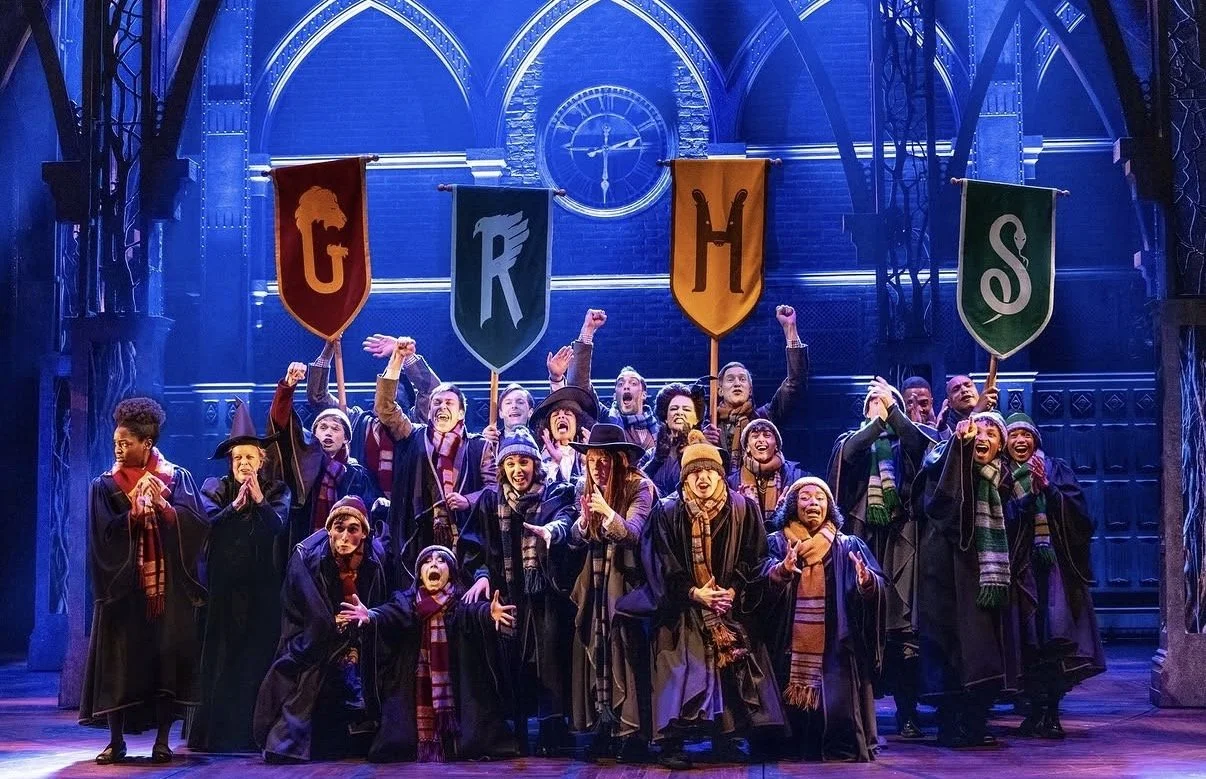 The cast of Harry Potter performing on stage, holding banners with the letters G, R, H, and S, dressed in Hogwarts robes and scarves, with a blue-lit castle-themed set in the background.