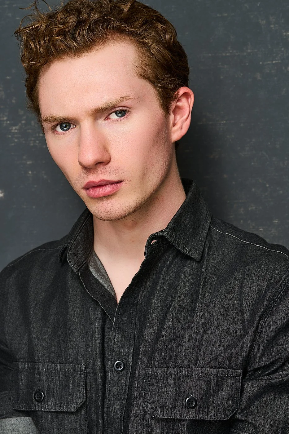 A young man with curly red hair and blue eyes, wearing a black denim shirt, standing against a dark textured background.