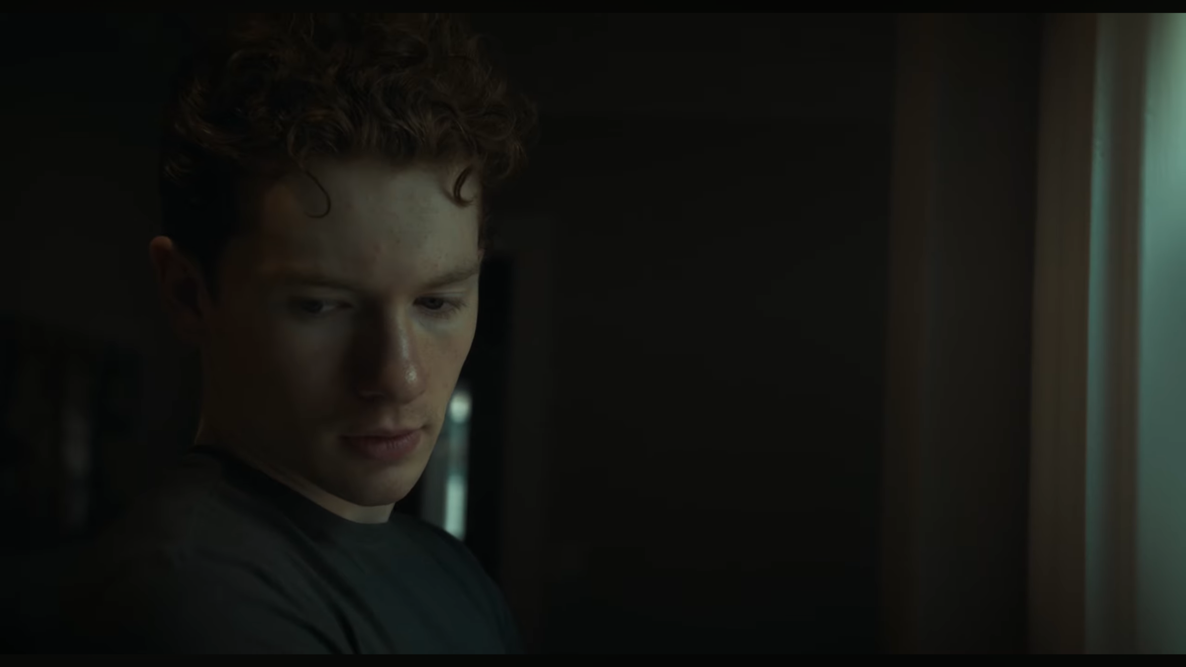 A young man with curly hair looking downward, illuminated by a soft light coming from the right side of the image, with a dark background.