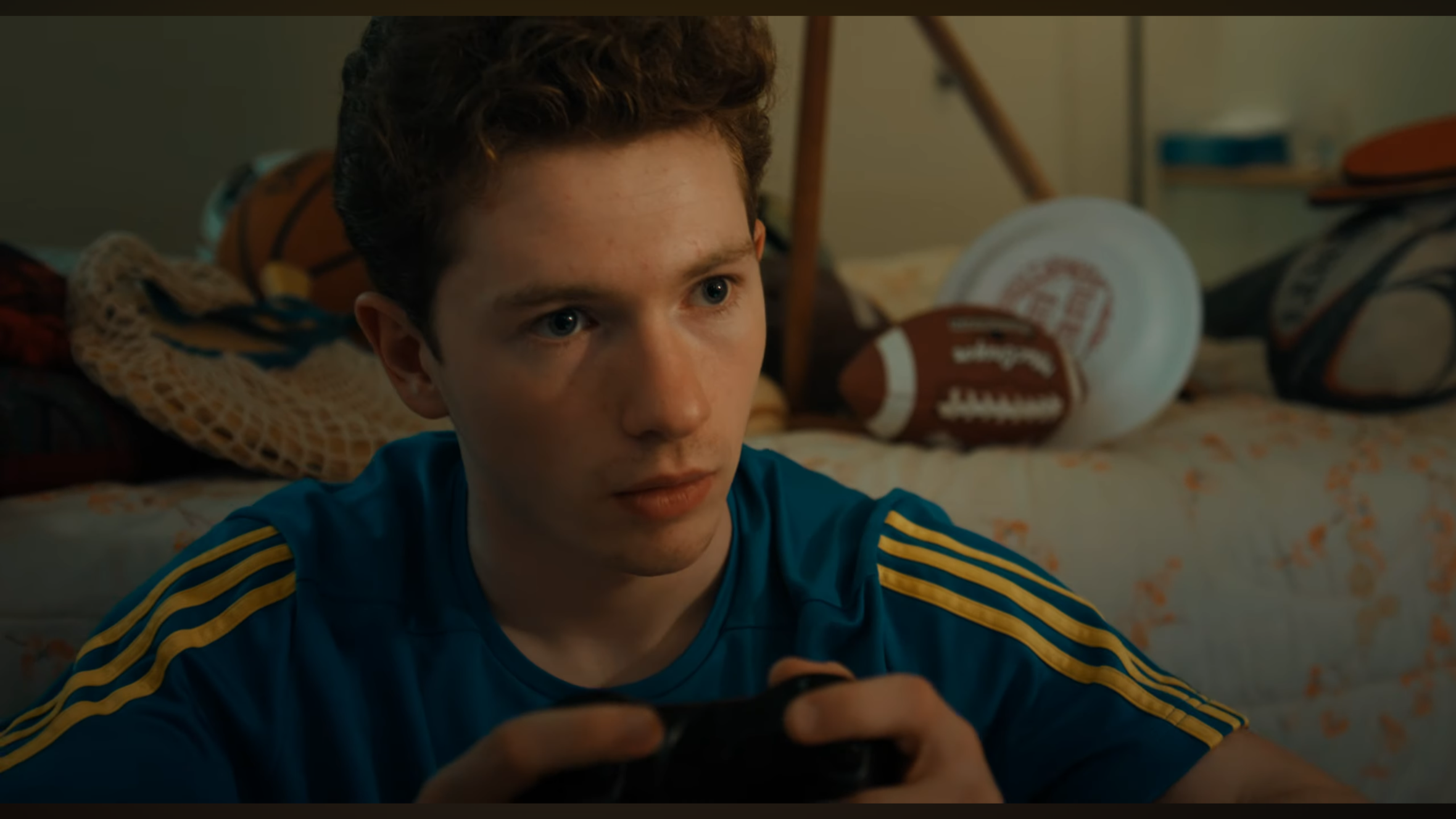 A teenage boy with curly hair and blue eyes, wearing a blue sports jersey with yellow stripes, sitting in a bedroom with sports equipment, including basketballs, footballs, and a baseball mitt, in the background.