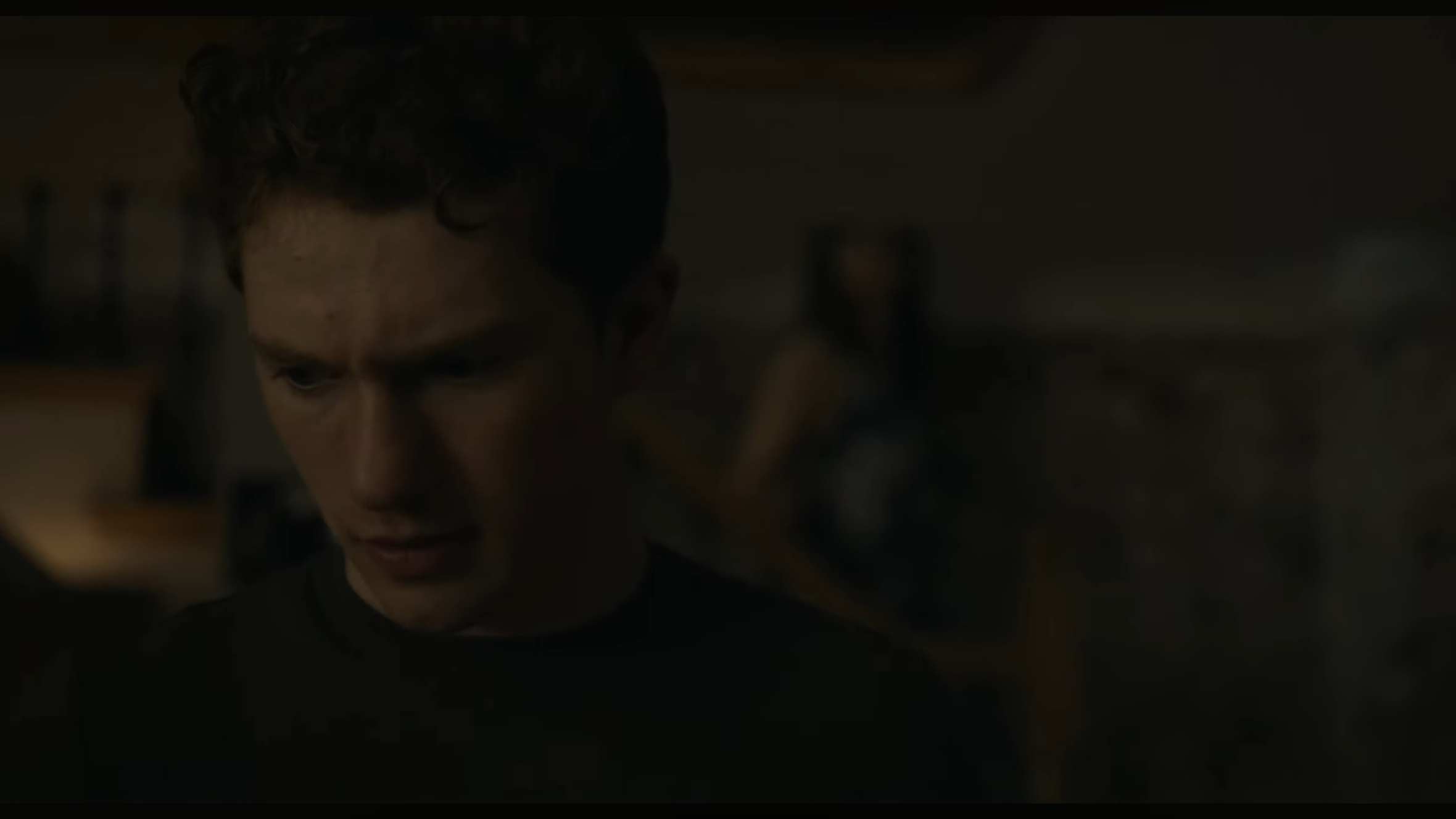 A young man with curly dark hair and a serious expression, looking downward, in a dimly lit room.