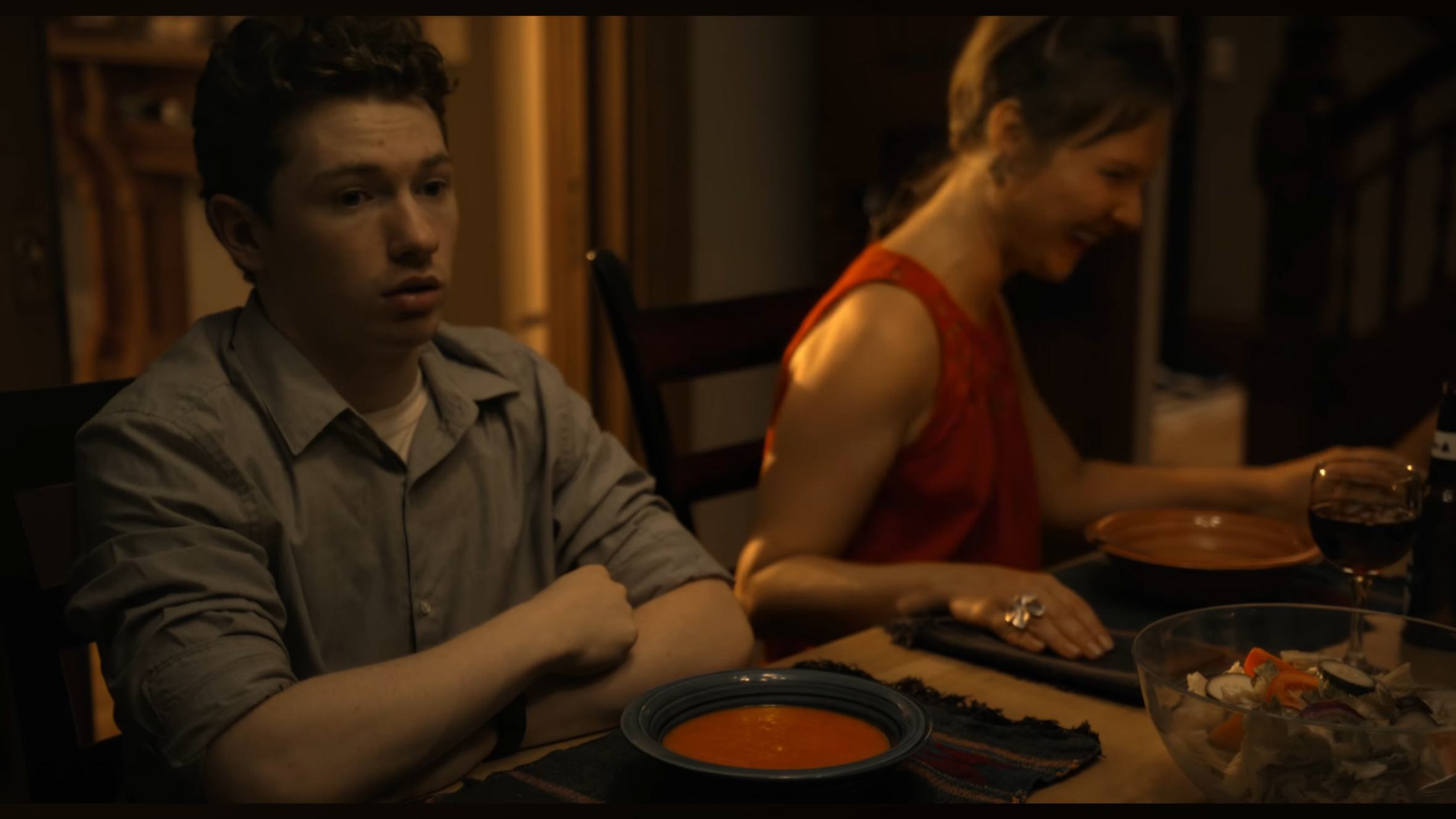A young man with dark, curly hair and a woman with brown hair styled in a braid sit at a dinner table with food and drinks, sharing a meal in a warmly lit room.