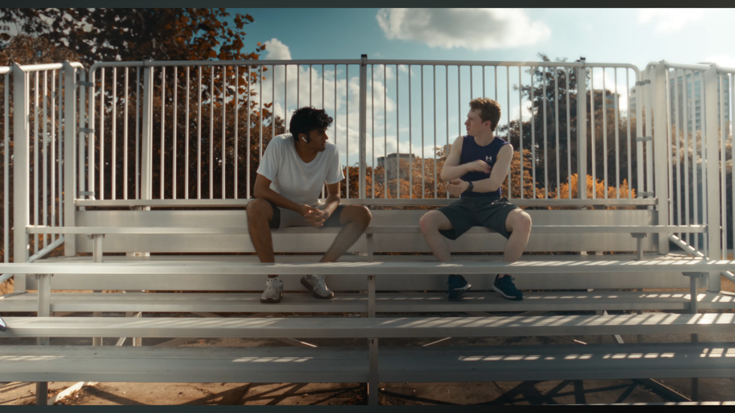 Two young men sitting on bleachers in a park, talking, with trees and clouds behind them.