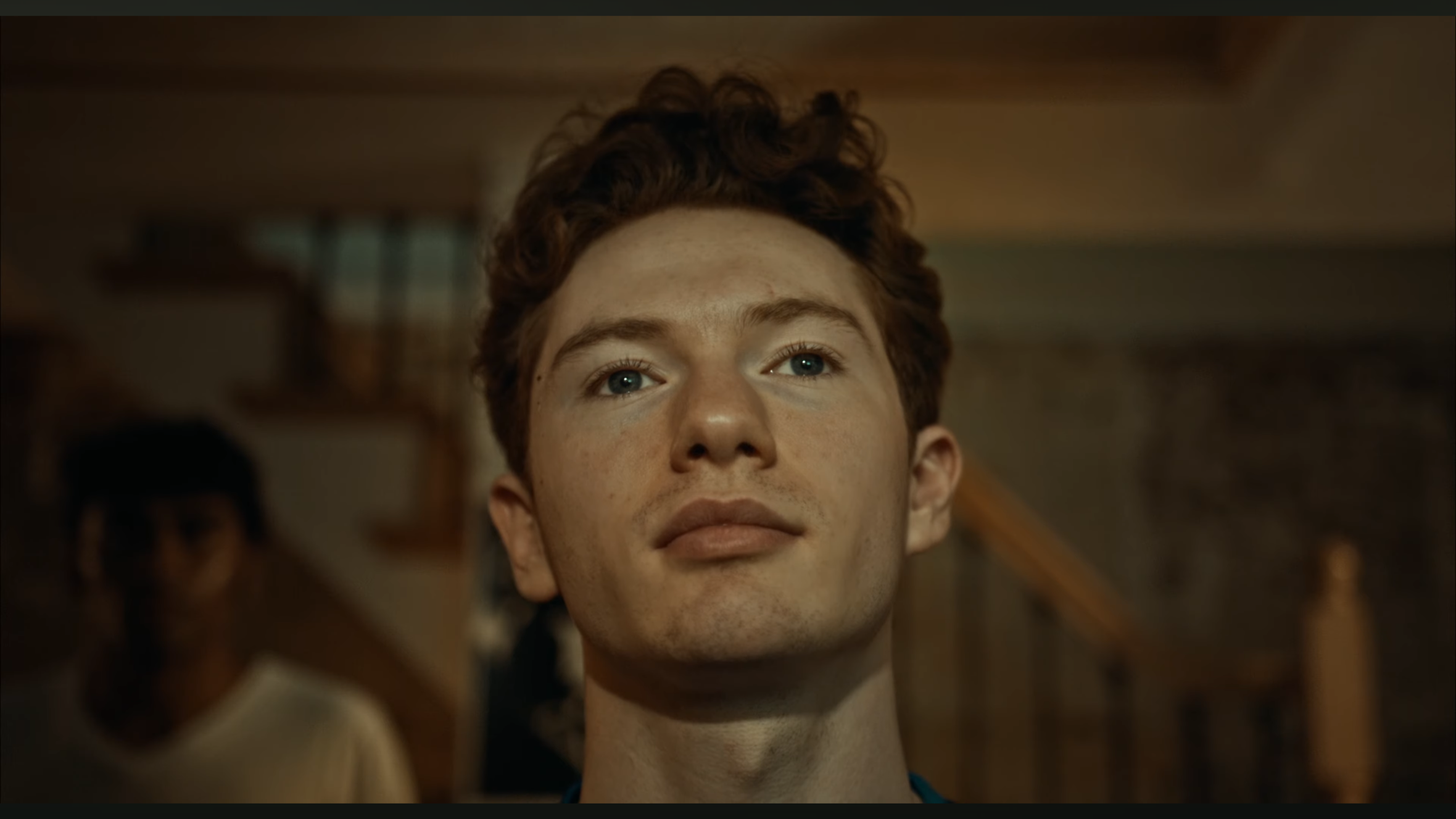 A young man with curly red hair and blue eyes looking up, with a serious expression, in an indoor setting.