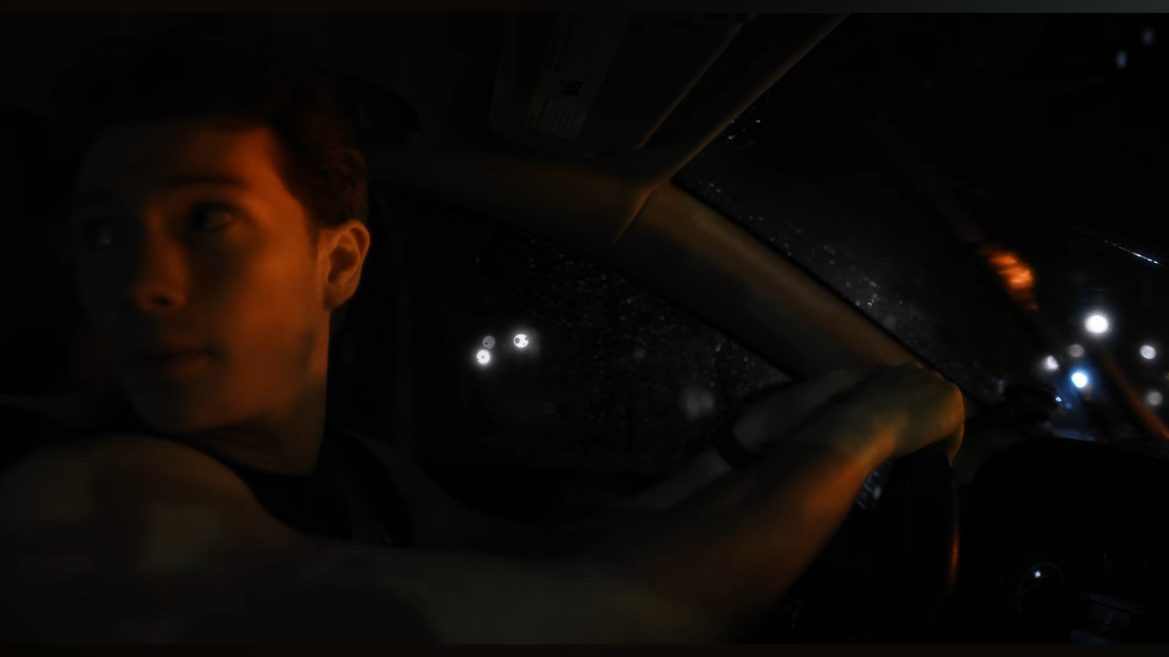 A man with short hair sitting in a car at night, looking to the side, with rain droplets on the windshield and city lights in the background.