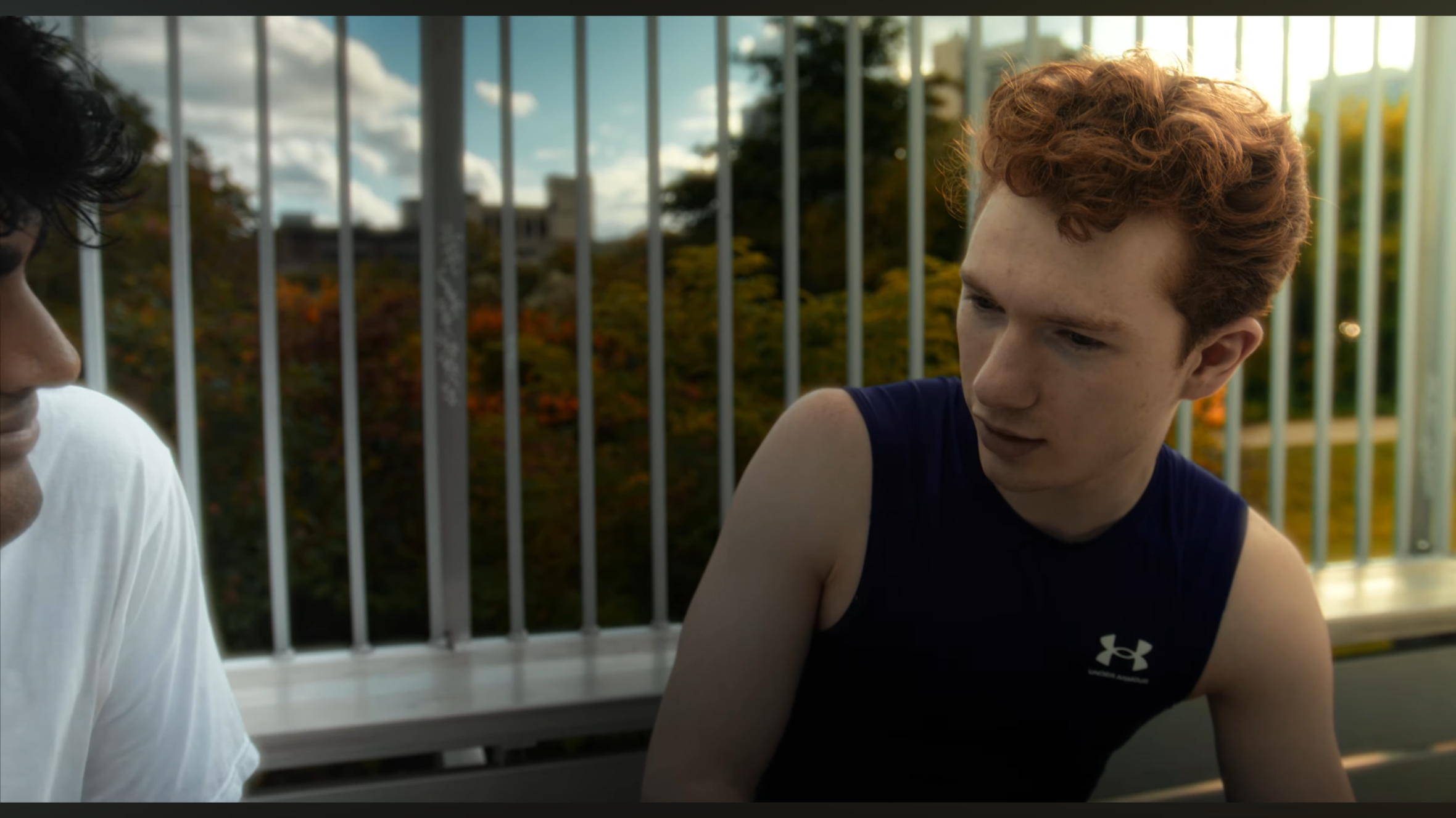 Two young men sitting outdoors near a white railing with trees and a cloudy sky in the background, engaged in conversation.
