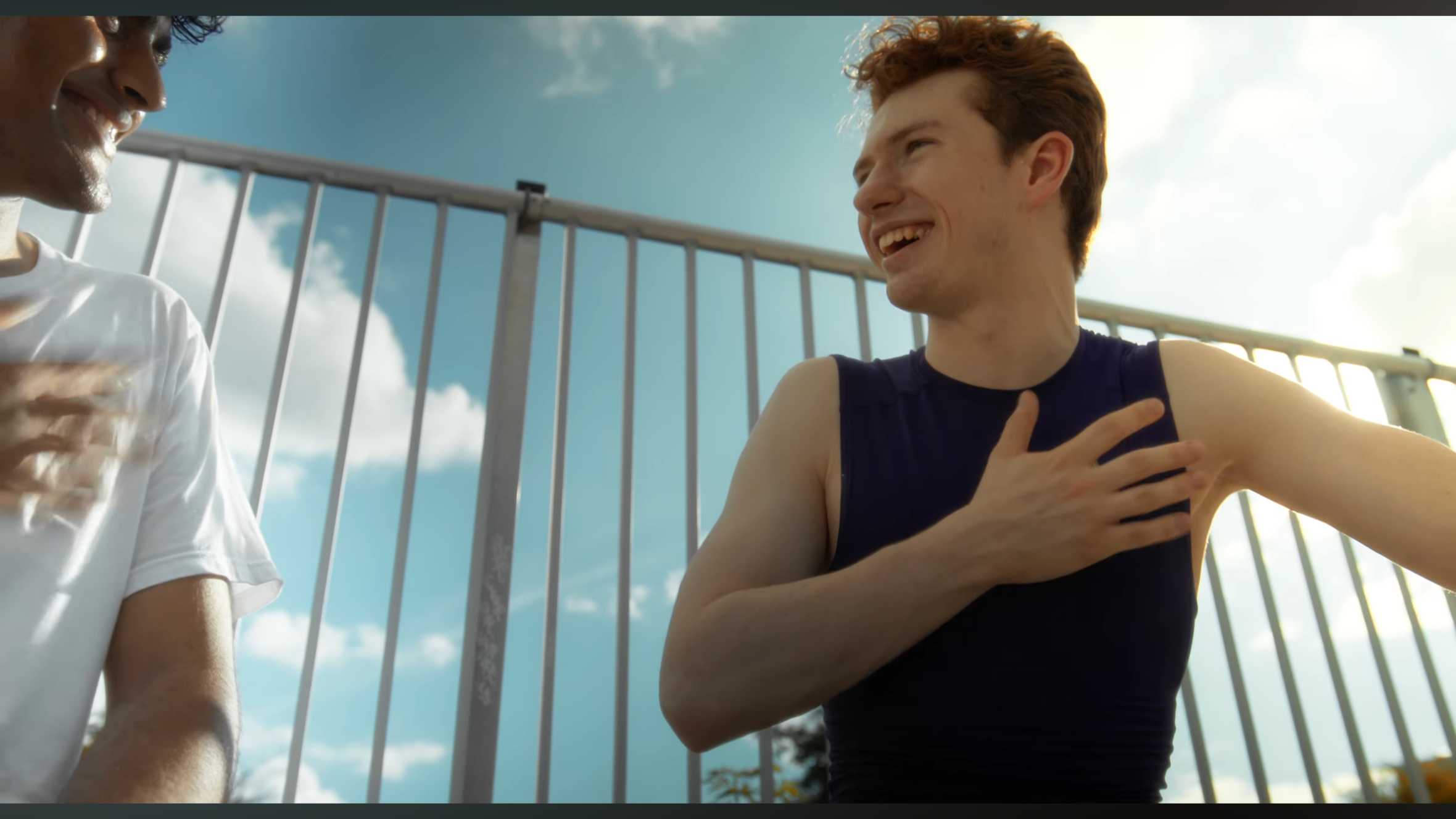 Two young men outdoors near a metal fence, smiling and laughing during daytime, with one touching his chest and the other rubbing his shoulder, under a partly cloudy sky.