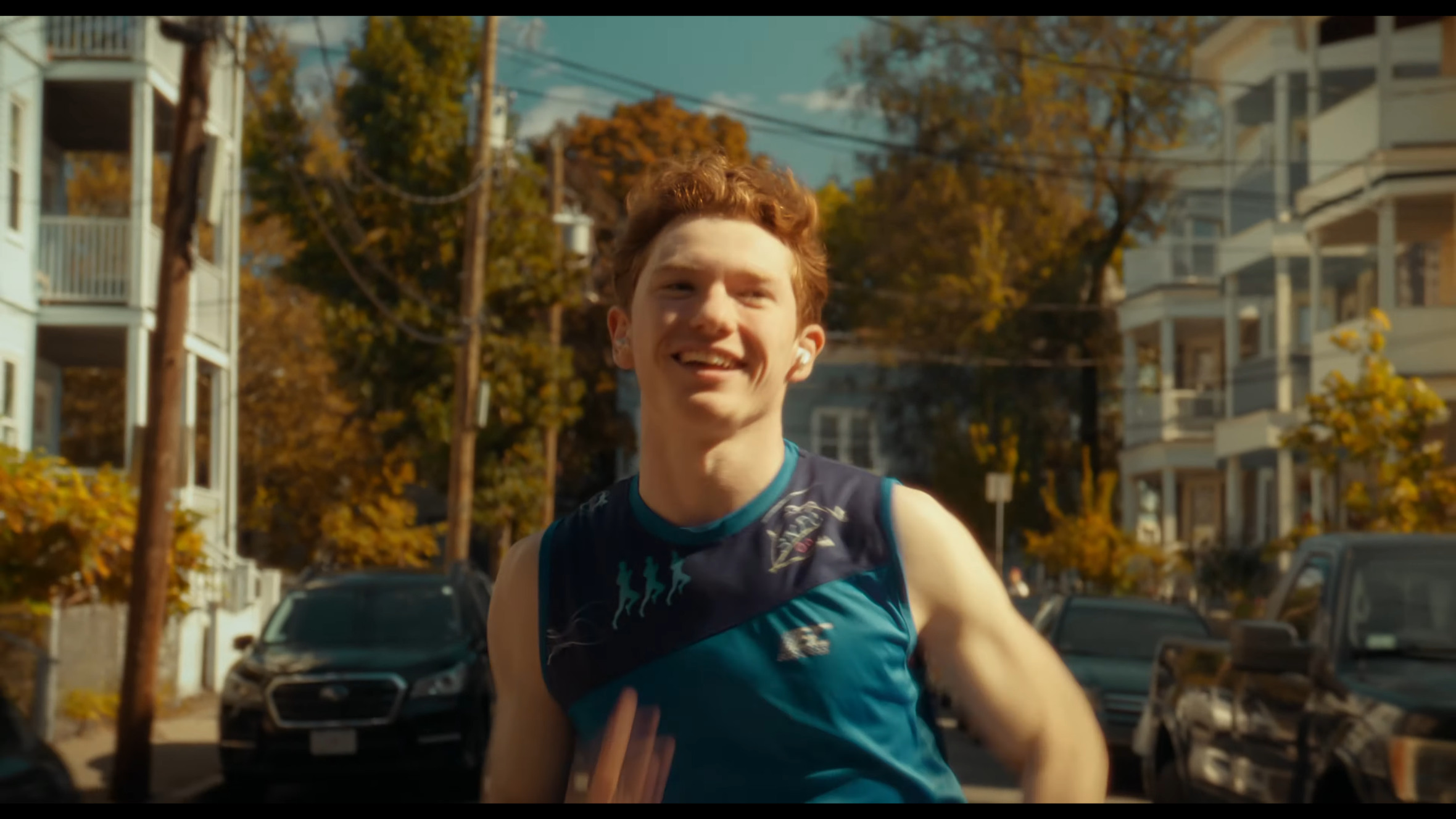 A young man with red hair smiling while running outdoors on a sunny day, with trees and buildings in the background.