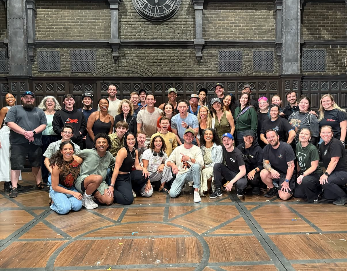 A large group of diverse people smiling and posing for a group photo indoors, with a brick wall and clock in the background and a wooden floor in the foreground.