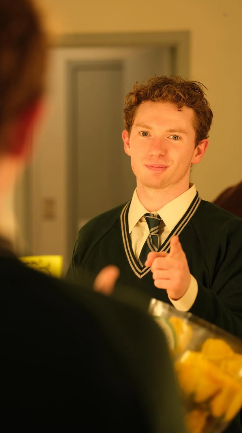 Young man in school uniform pointing at the camera while smiling, seen through a mirror, with a bowl of snacks in the foreground.