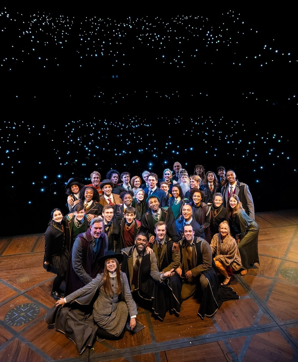 A large group of people dressed in Hogwarts uniforms, some with robes, ties, and hats, posing together on stage in front of a black background with twinkling lights.