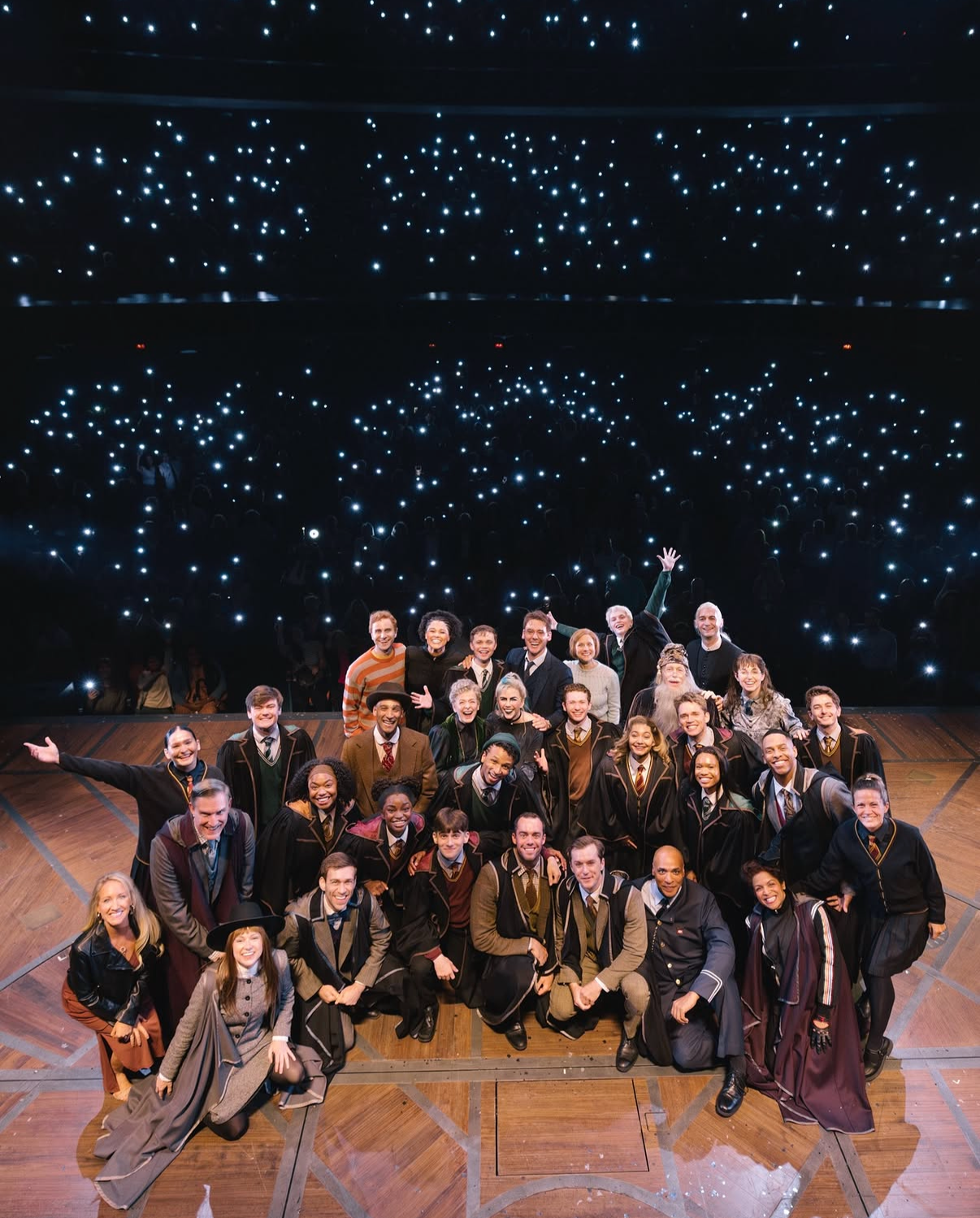 A group of diverse people in graduation gowns posing on stage with an audience and starry ceiling in the background.