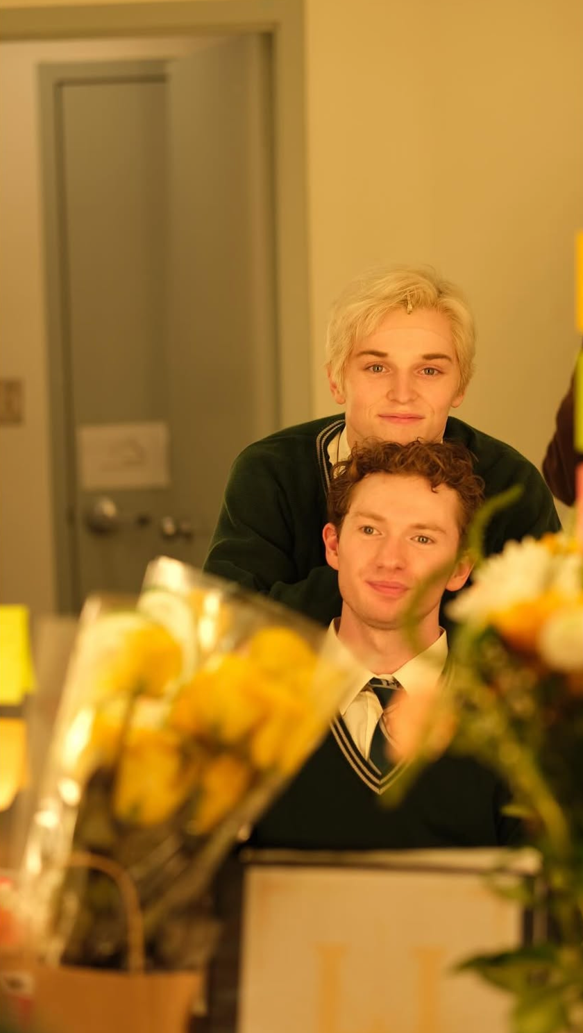 Two young men with blond and curly brown hair, smiling, are looking into a mirror in a room with a yellowish tint. Flowers and a cardboard box are in the foreground.