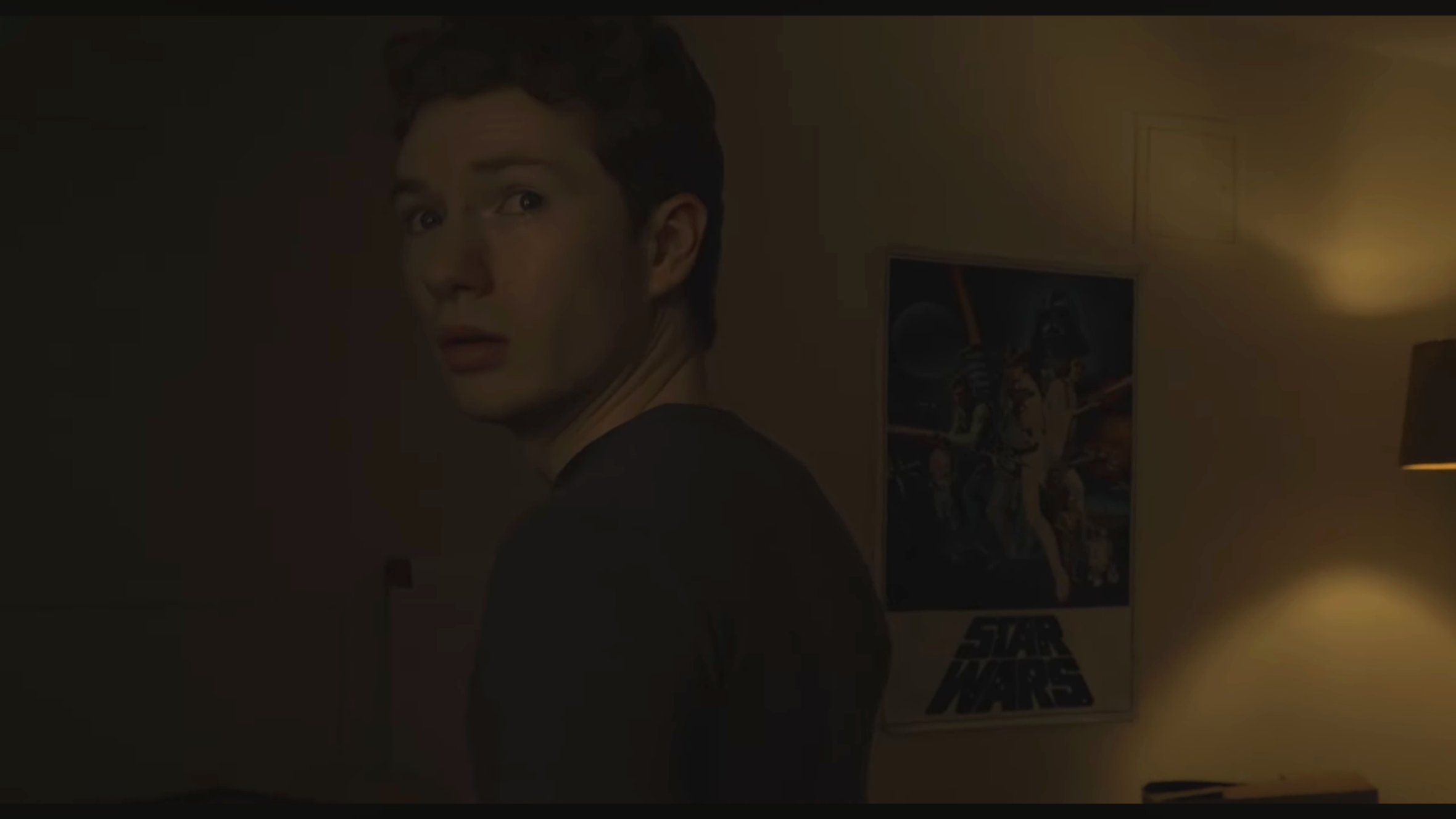 A person with dark hair looking over their shoulder in a dimly lit room. In the background, there is a Star Wars poster on the wall and a lamp casting warm light.