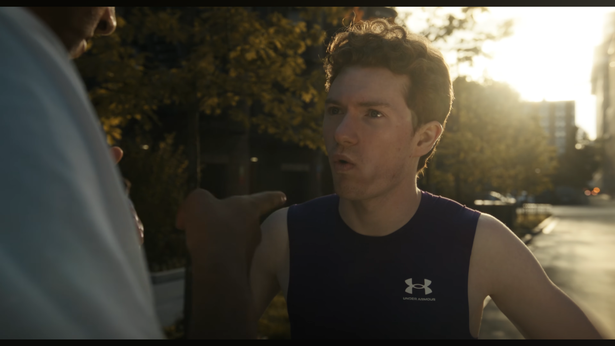 A young man with curly hair and a serious expression is being confronted by another person during what appears to be an argument or confrontation outdoors at sunset.