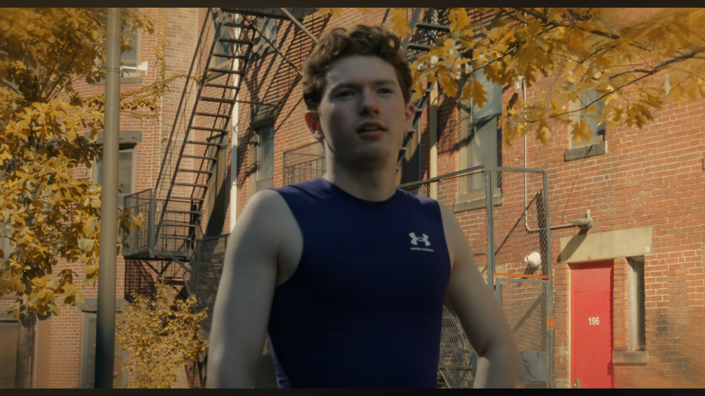 A young man with curly hair wearing a navy blue Under Armour athletic shirt outdoors on a sunny day, with a brick building, fire escape, and autumn-colored trees in the background.