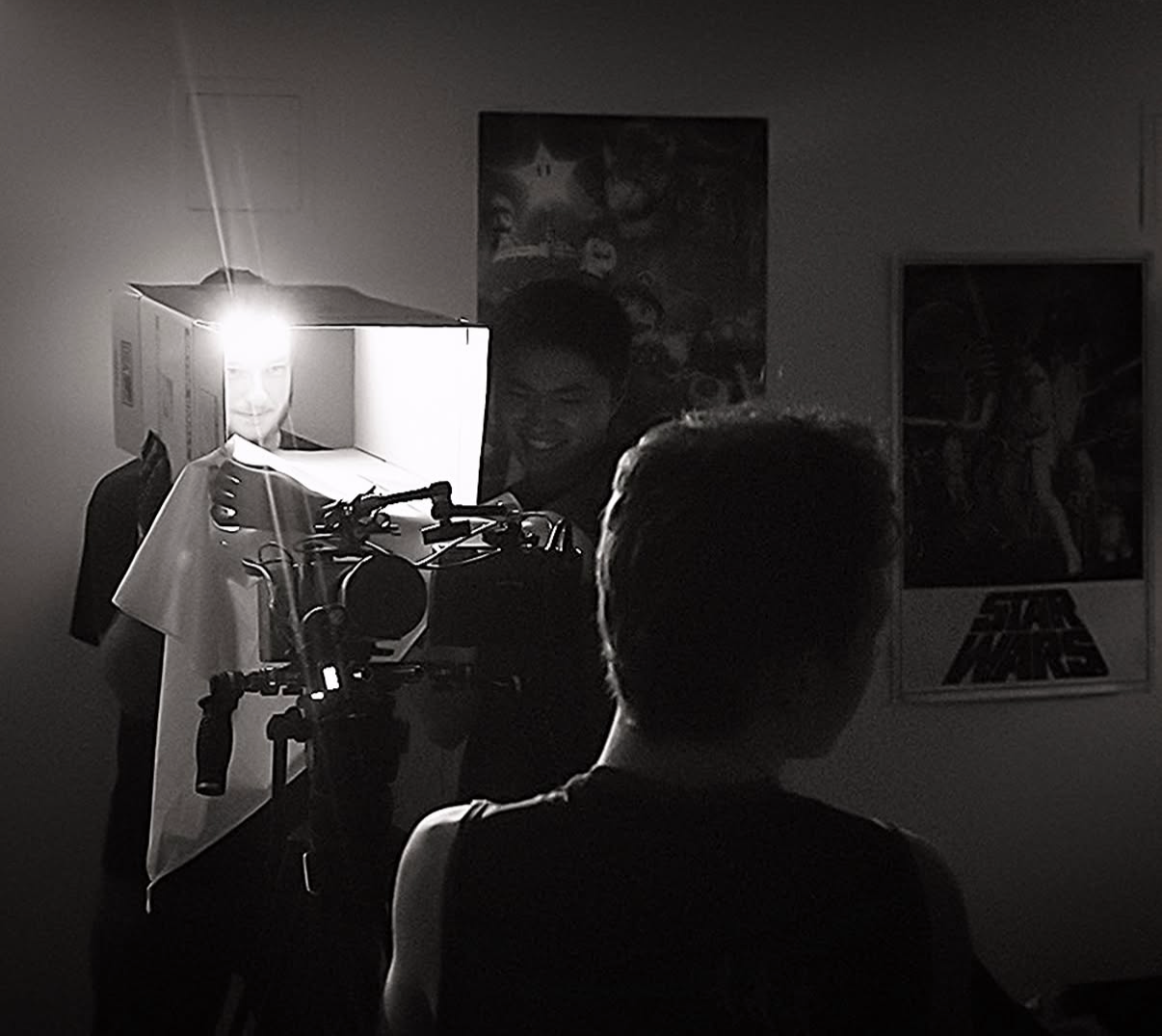 A person filming a figure dressed in a tuxedo with a white shirt, standing behind a device with a bright light on a dark room, with posters on the wall in the background.