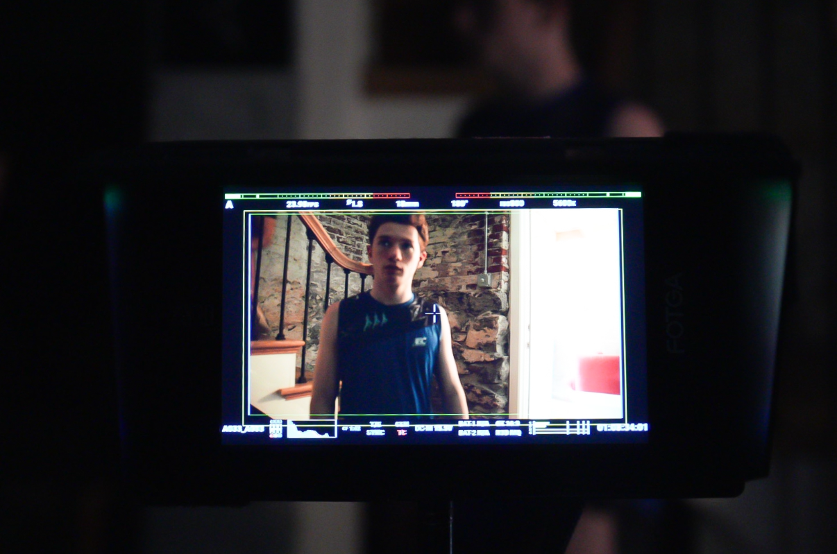 A person wearing a sleeveless shirt being filmed or photographed, with a stone wall and a staircase in the background.