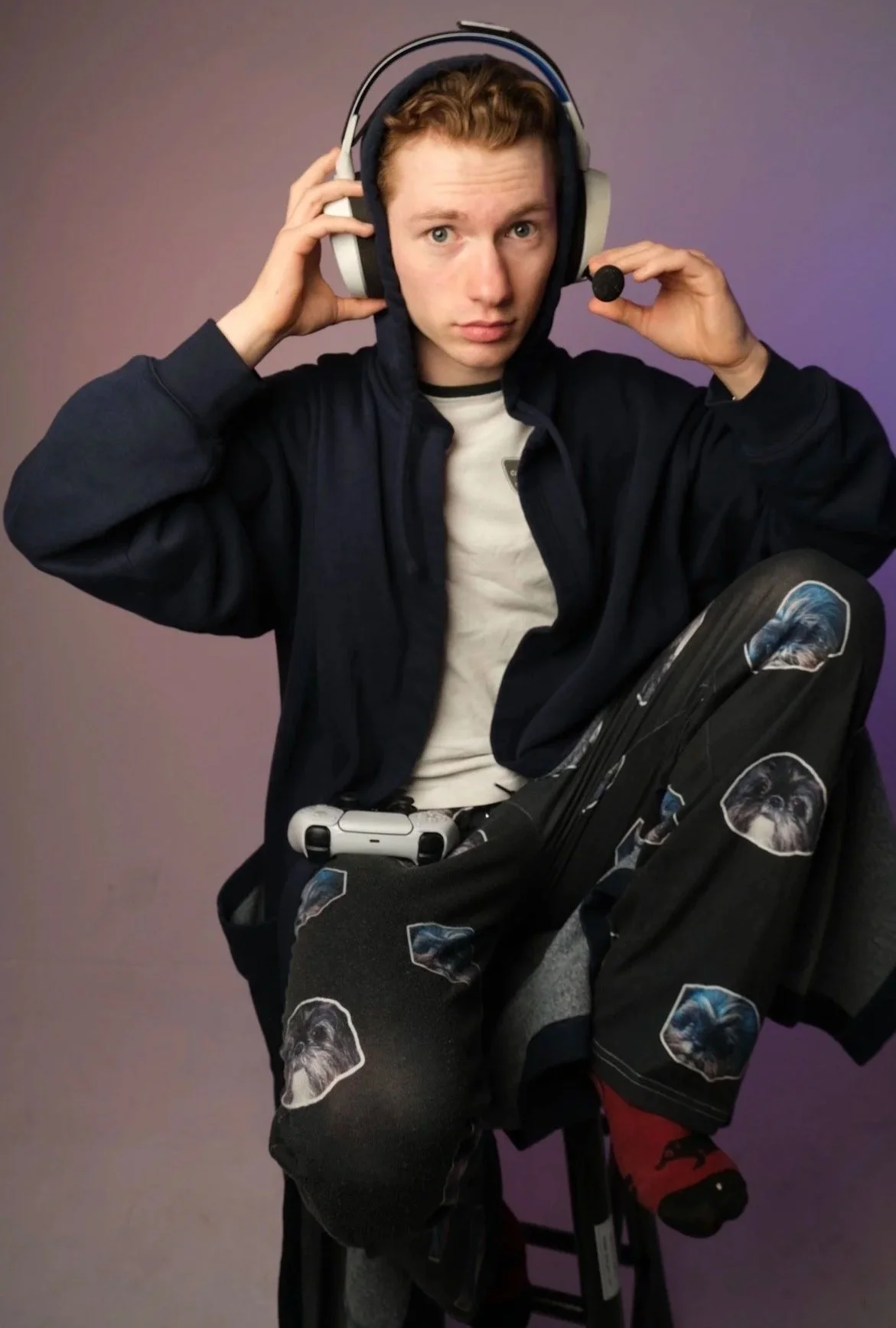 A young man with reddish curly hair is wearing headphones, a black hoodie, and a white T-shirt. He appears to be adjusting his headphones while sitting in front of a purple background, with a game controller resting on his lap and matching pants featuring dog faces.