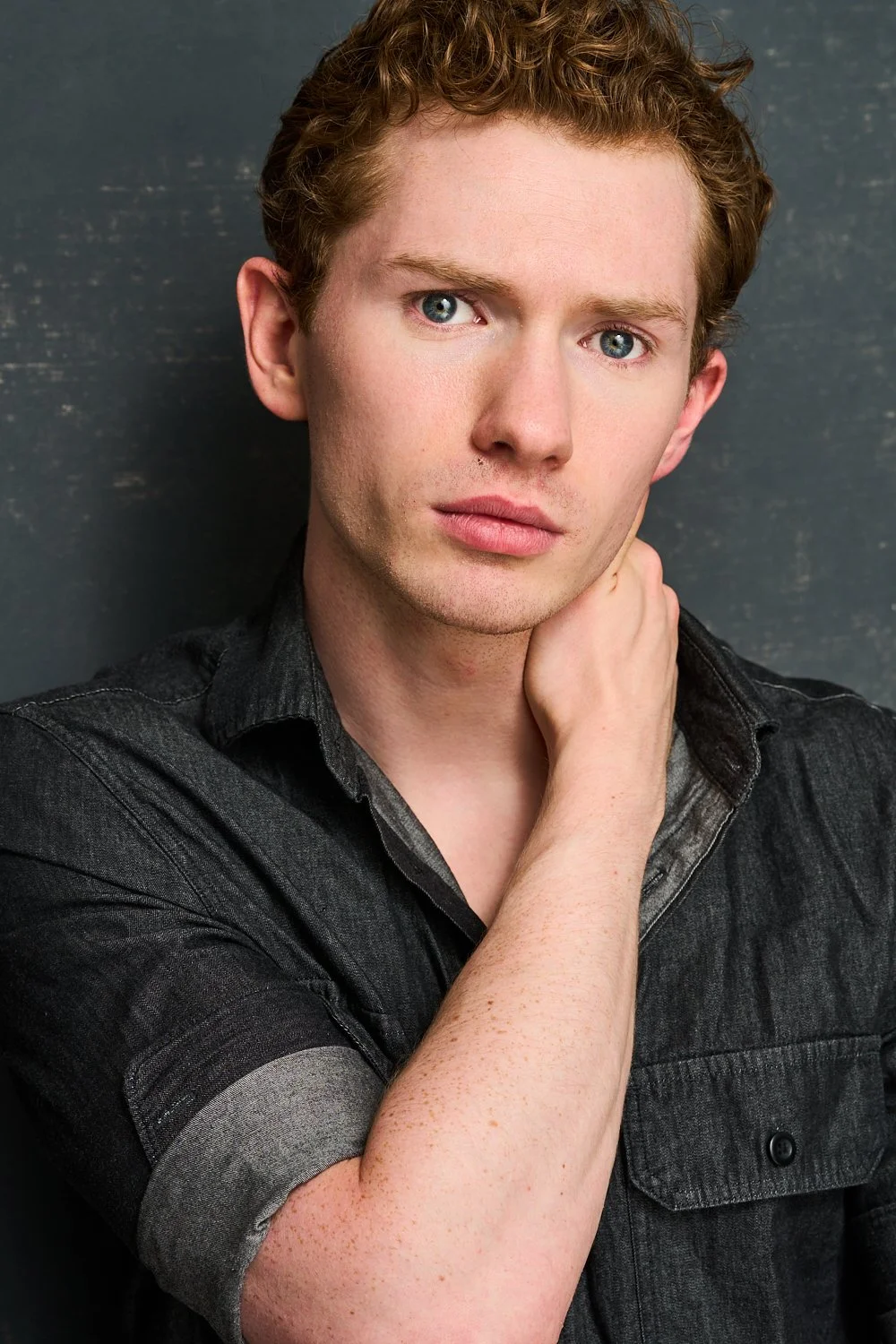 Portrait of a young man with red hair, blue eyes, wearing a black button-up shirt, standing against a dark background.