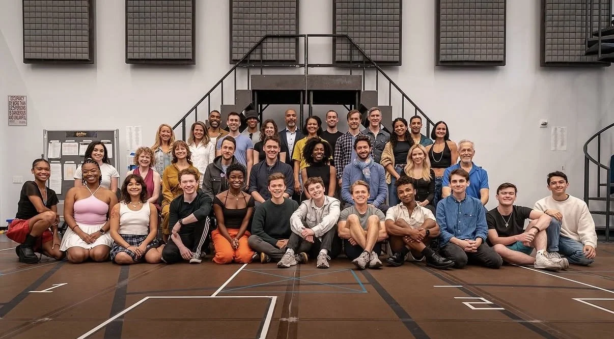 A large group of diverse people, including children and adults, posing for a group photo in a gymnasium or community center with basketball court markings on the floor.
