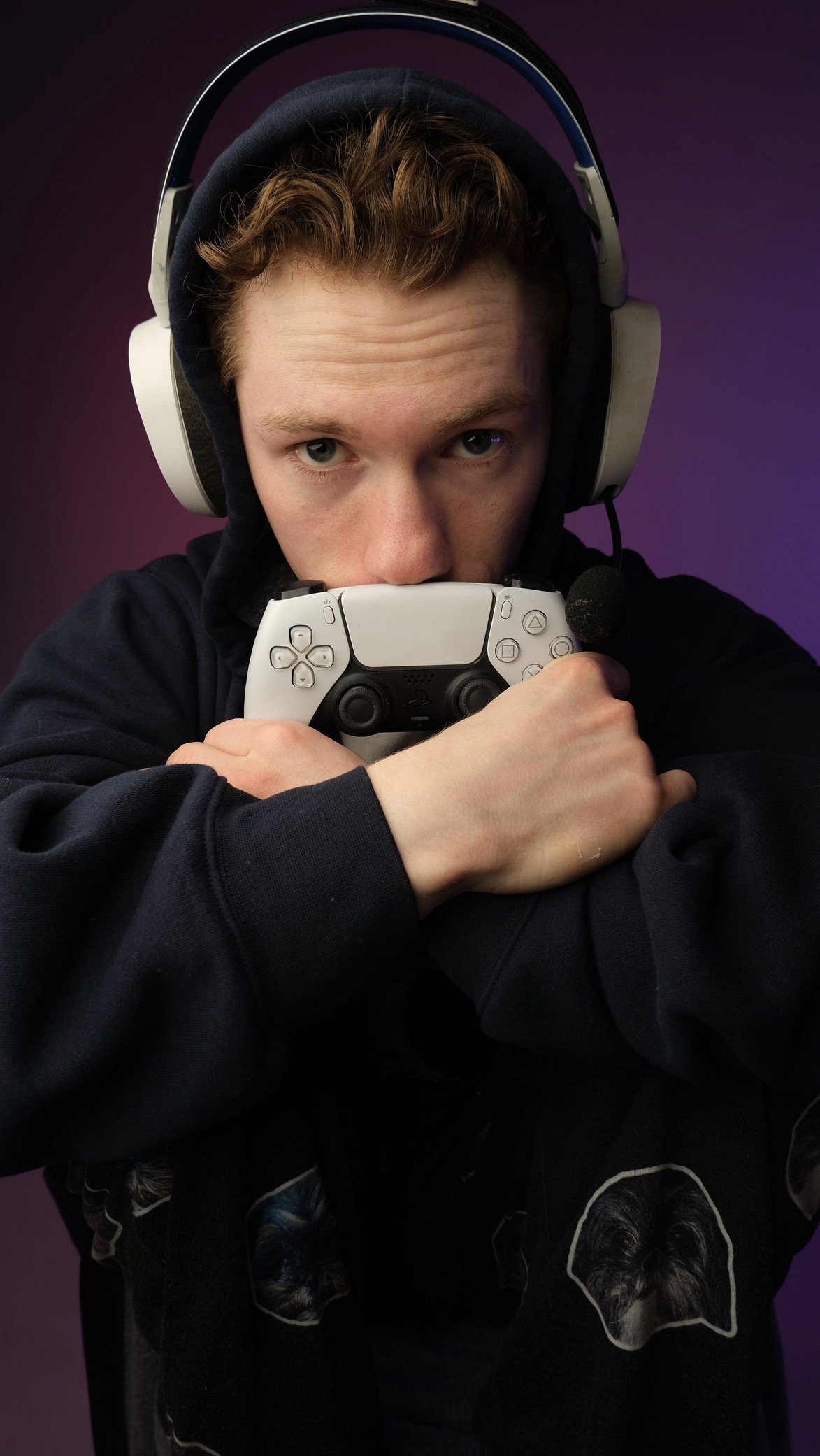 A young man with curly brown hair wearing a black hoodie and large over-ear headphones, holding a white PlayStation controller, with a purple background.