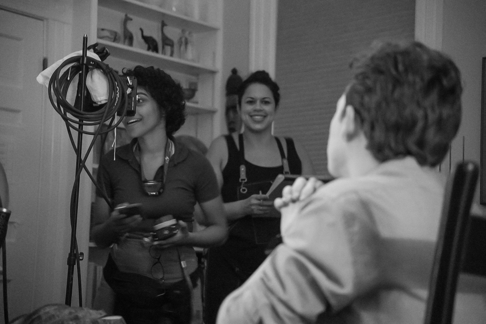 Two women standing and smiling, facing a man seated with his back to the camera, in a room with shelves and decorations in the background, black and white photo.