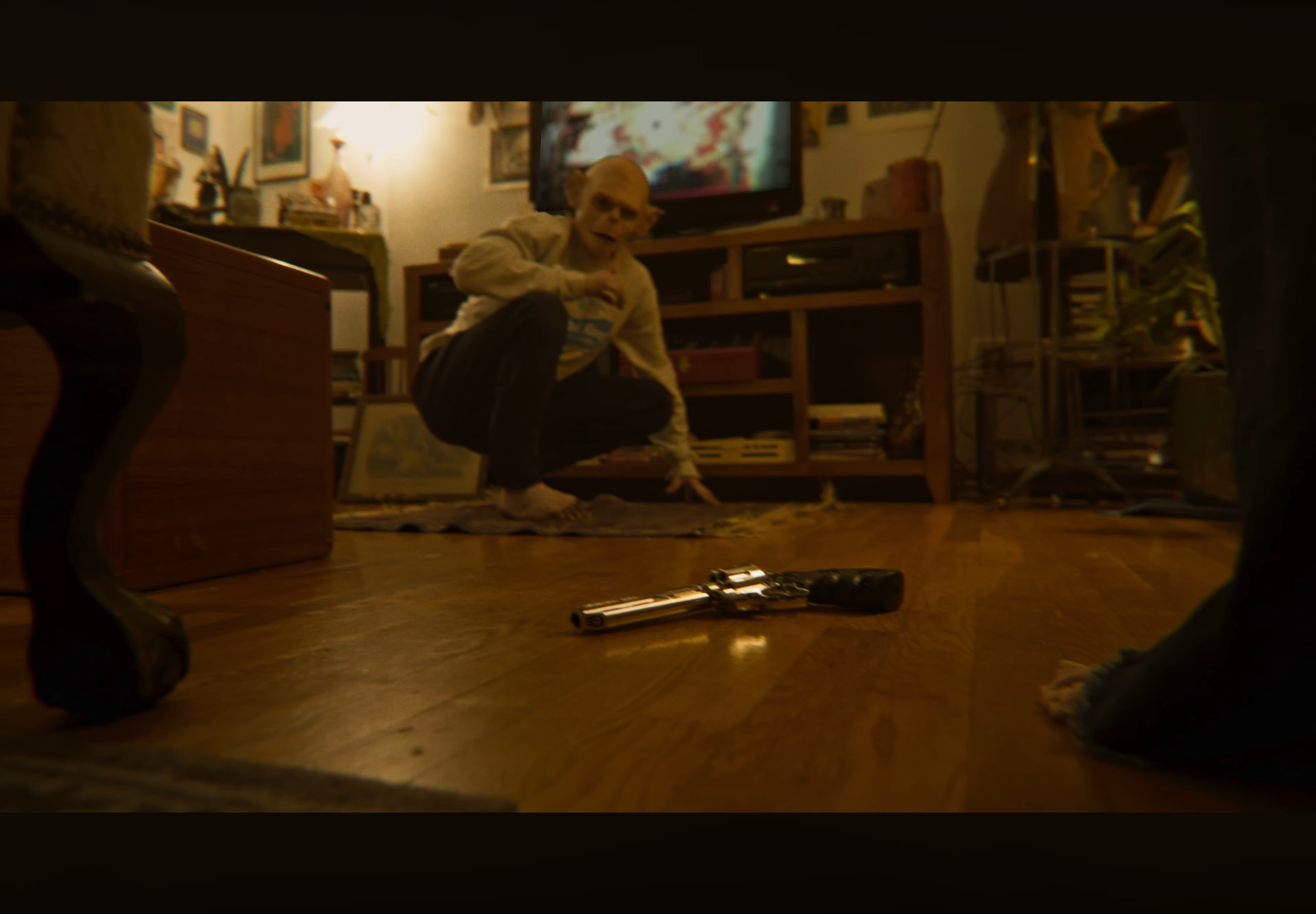 A person dressed like a skeleton is kneeling on the floor in a dimly lit room, with a TV and bookshelves in the background. A gun is lying on the floor in the foreground.