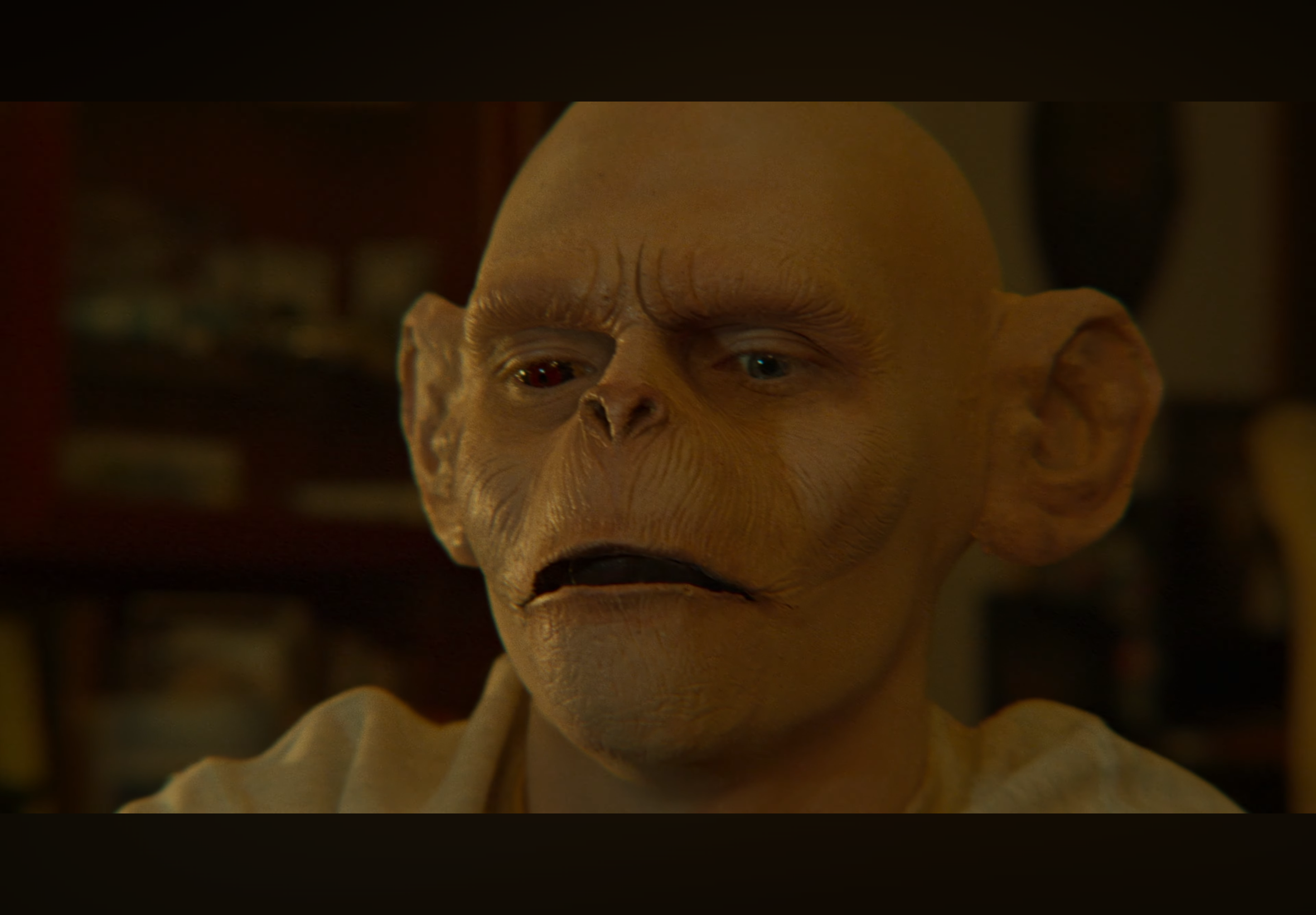 Close-up of a humanoid creature with a large, smooth, bald head, textured skin, small ears, and one eye with a red iris, looking upward.