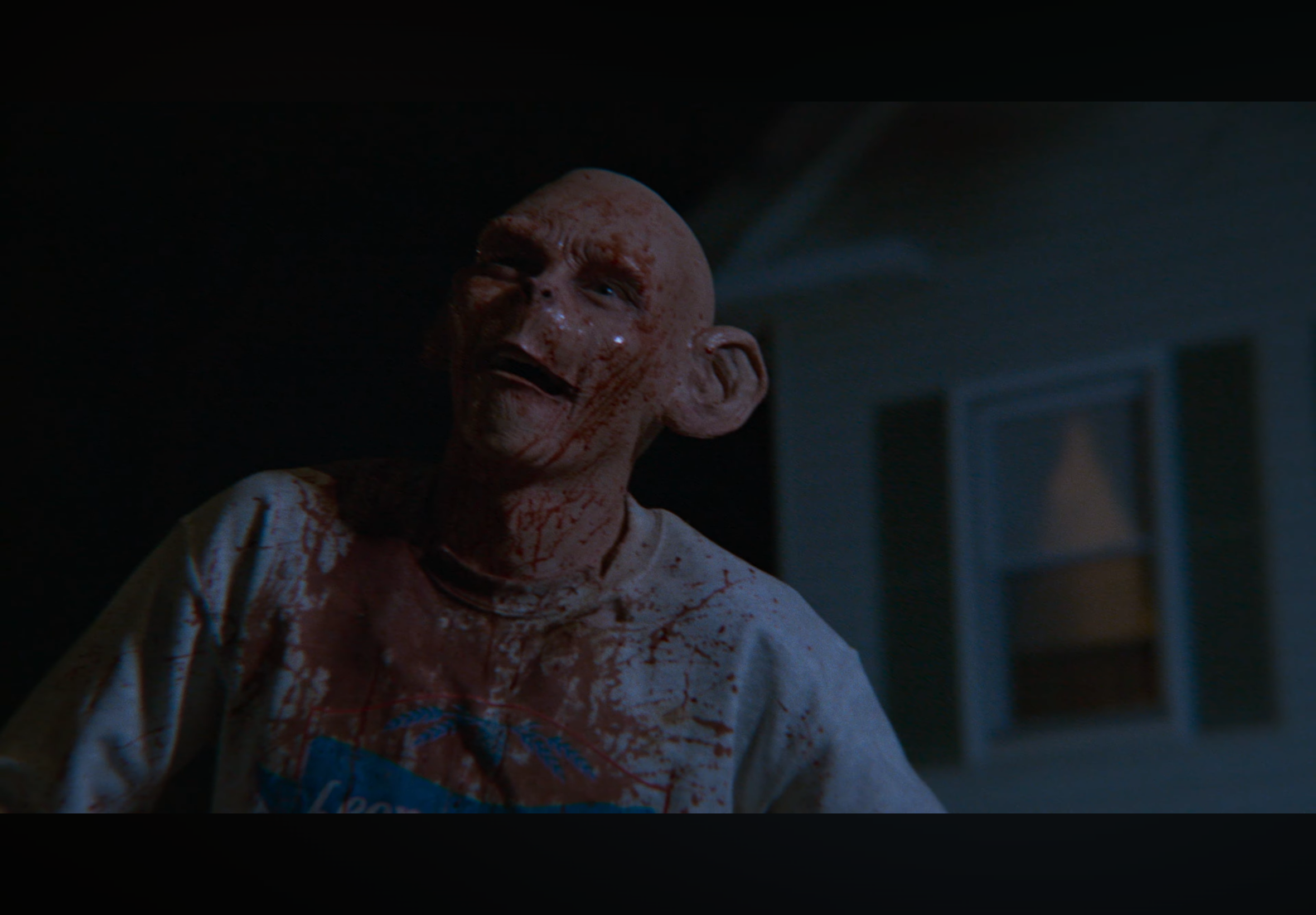 A humanoid creature with a large bald head and dark eyes, covered in blood, standing in a dimly lit room.