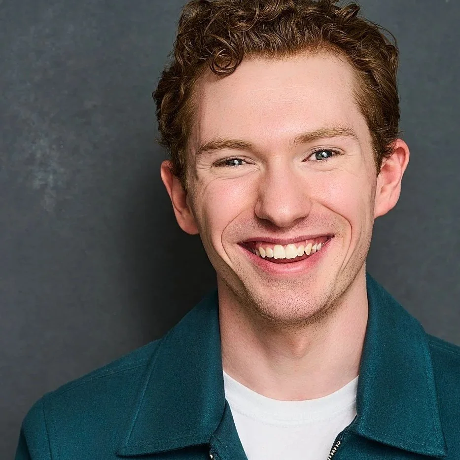 A smiling Zach Norton with curly red hair, fair skin, wearing a teal jacket over a white shirt, standing against a gray background.