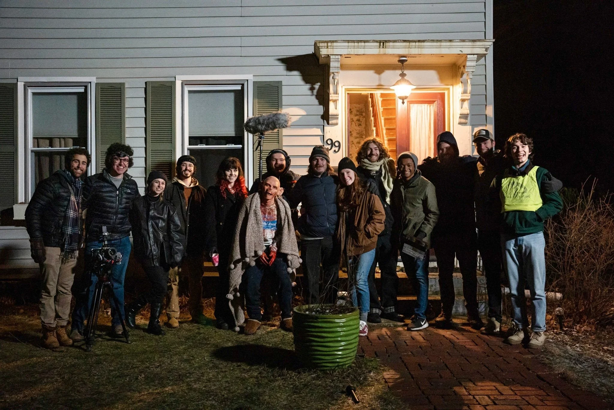 Group of 13 people standing outside a house at night, smiling for the camera, with a film crew member and equipment visible. The house has blue siding, green shutters, and a lit porch with a staircase visible inside.