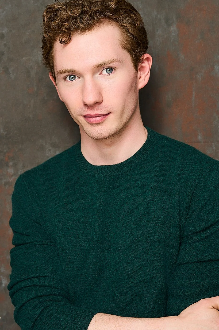 Portrait of Zach Norton with curly light brown hair, blue eyes, and fair skin, wearing a dark green sweater, posed against a textured brown background.