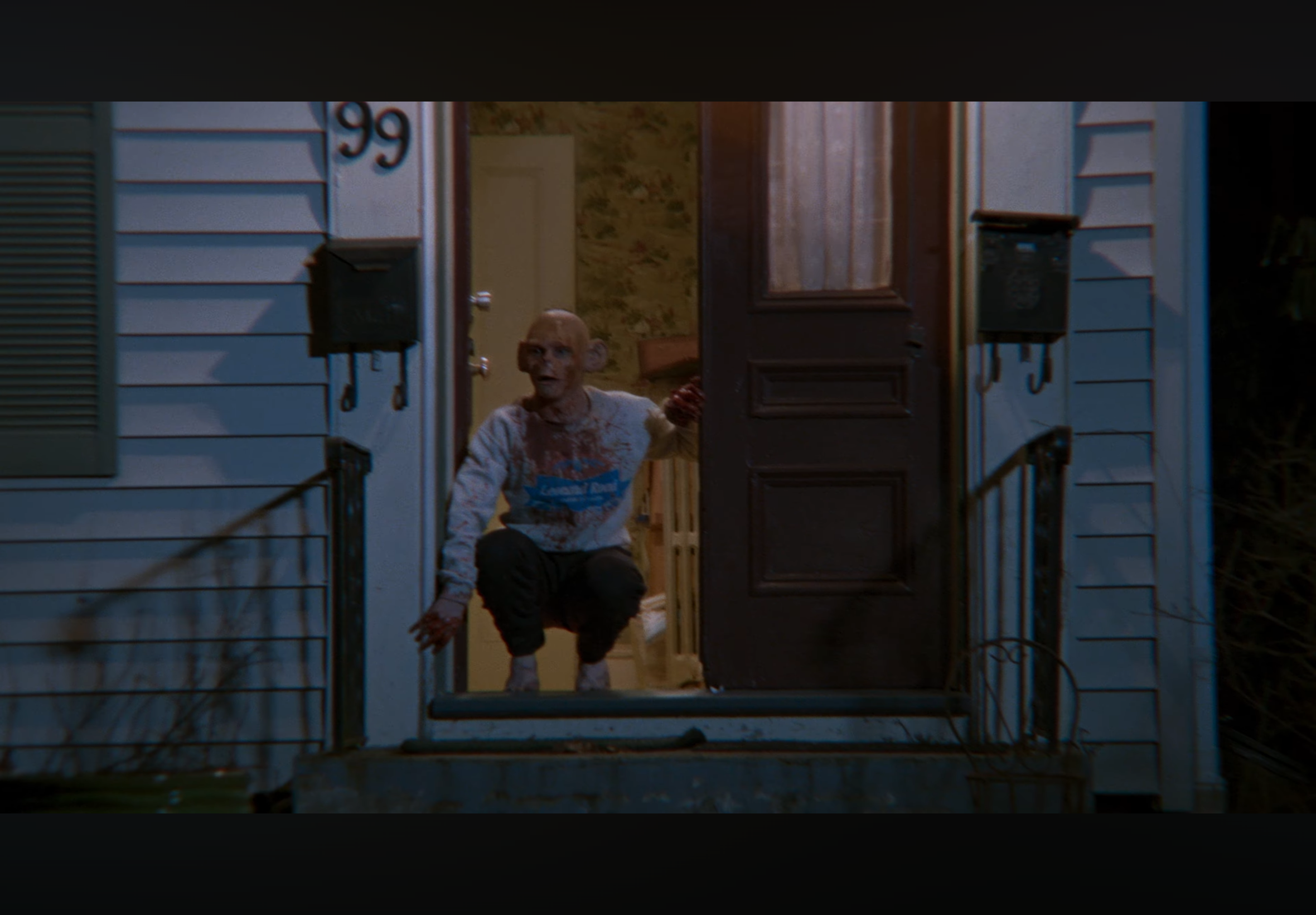 A zombie-like figure with a bald head, gaunt face, bloodstained white shirt, and black pants crawls out of a house at night. The person is reaching towards the camera with blood on their hands and face, standing at the open front door of a house with