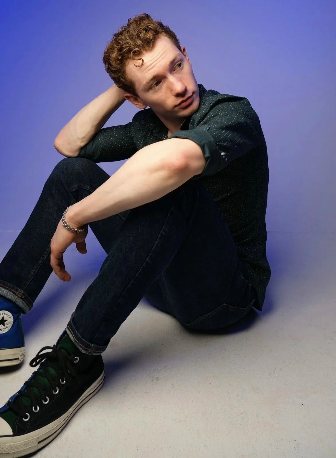A young man with curly red hair sitting on the floor against a purple background, wearing a dark button-up shirt, jeans, and Converse sneakers, looking thoughtfully to the side.