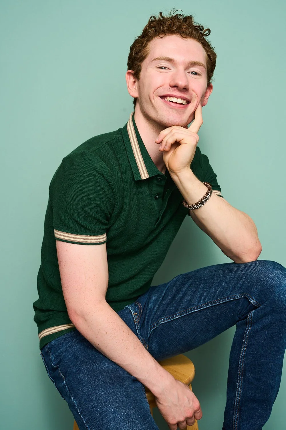 A young man with curly hair, wearing a green polo shirt with stripe details on the collar and sleeve cuffs, smiling thoughtfully while sitting on a stool against a plain light green background.