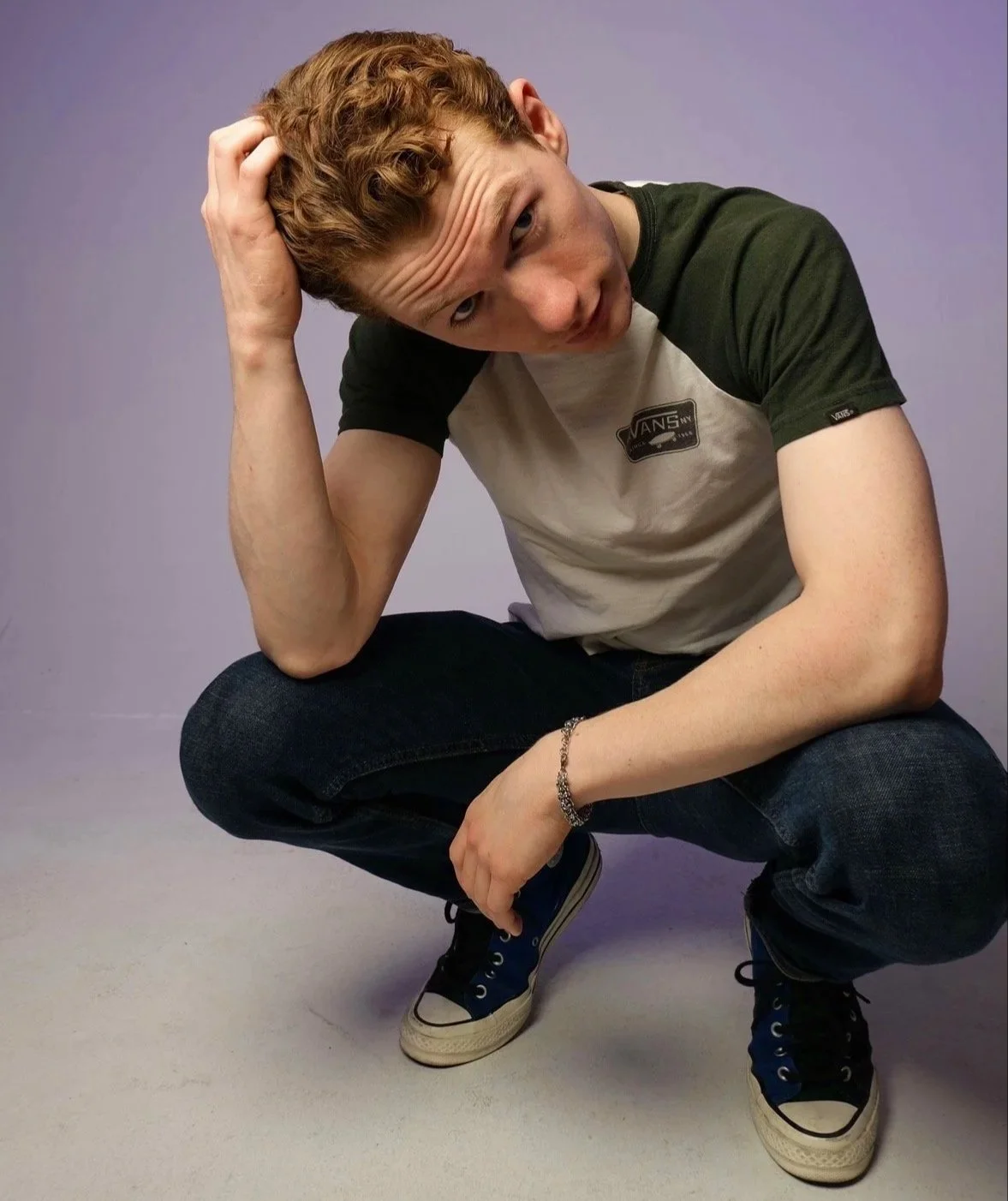 A young man with curly red hair crouches with one arm resting on his knee and his hand in his hair, wearing a beige and green Vans t-shirt, dark jeans, and blue sneakers, against a plain purple background.